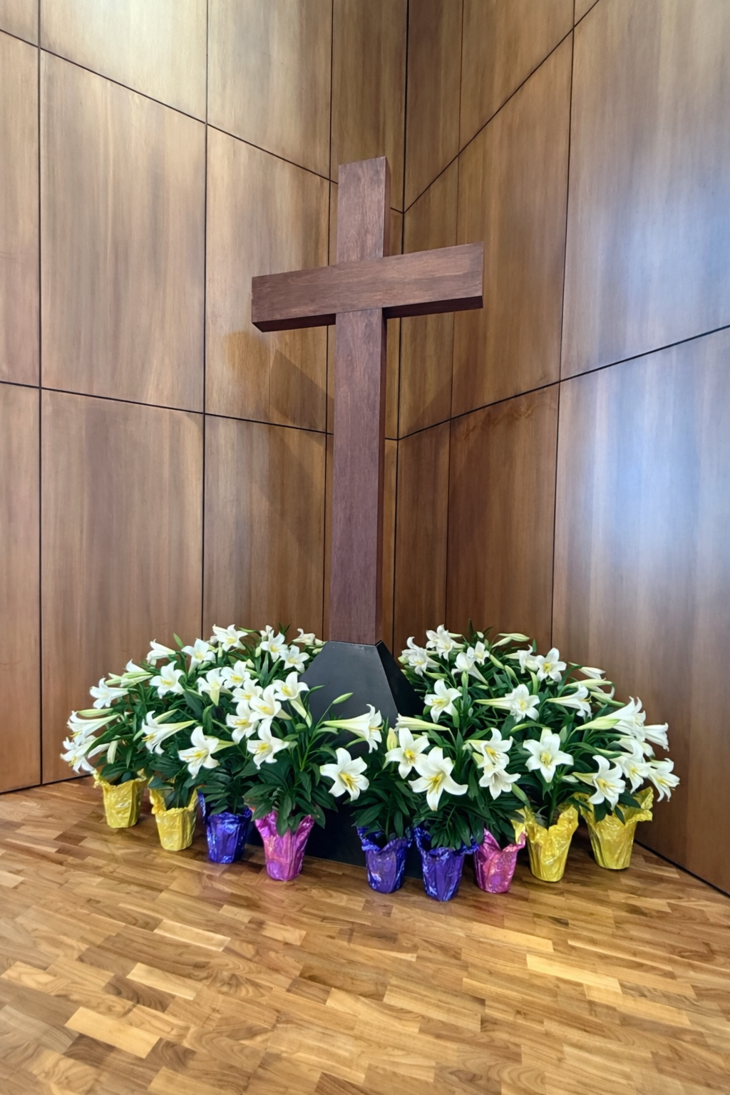A wooden cross inside a wood-paneled room, with Easter lilies around the base