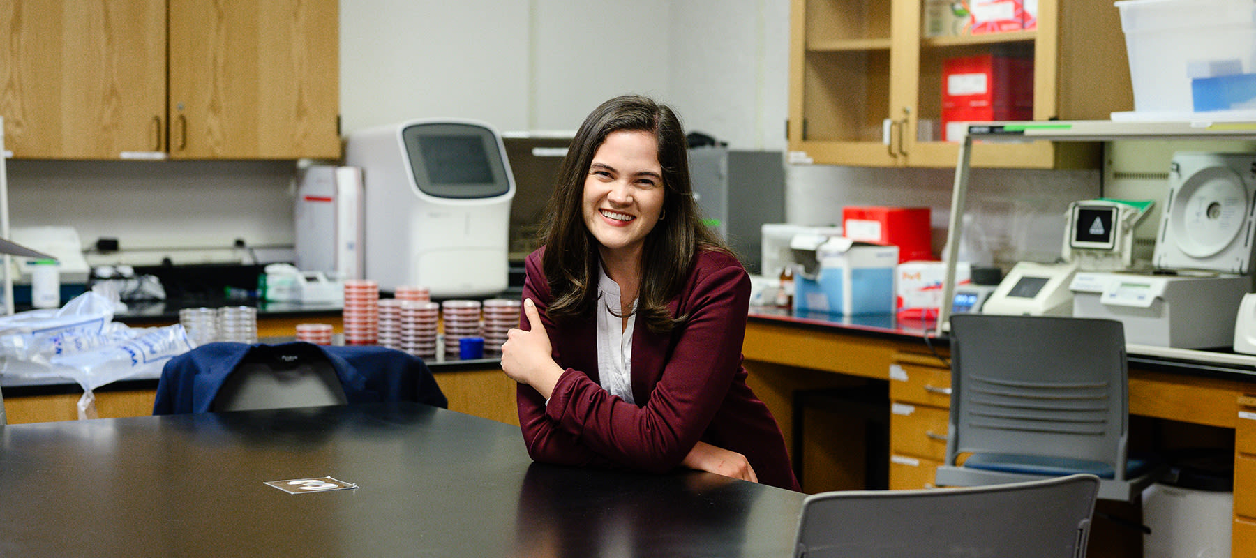 Sierra Rose wearing a suit jacket, leaning on a table and smiling in a CCU science lab