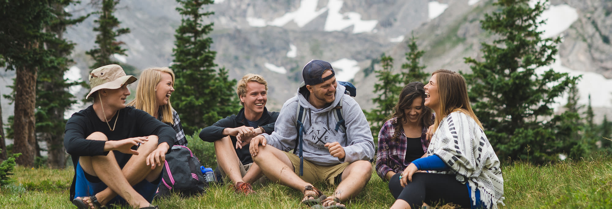 Students sitting outdoors in a circle, talking and laughing, with the Rocky Mountains visible in the background.