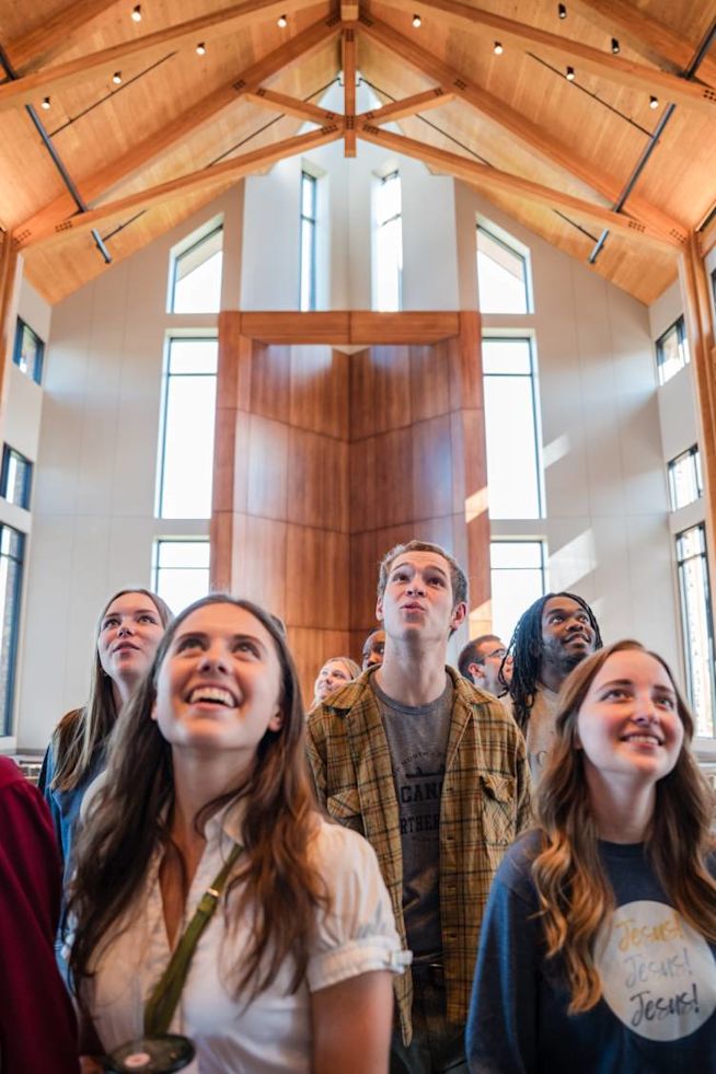 A group of college students stand inside the Considine Chapel with wooden beams and tall windows, looking upward with expressions of awe