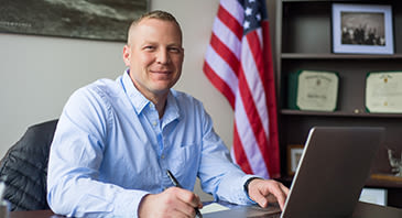 Smiling military student works at laptop, US flag seen in the background