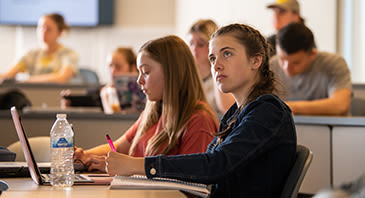 A classroom of students sitting at desks, looking forward attentively and taking notes.