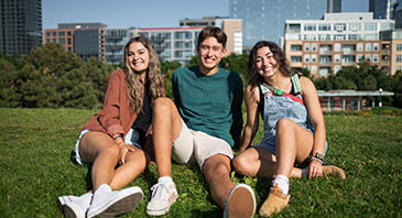 Three students sprawl on the grass, with the Denver skyline behind them.