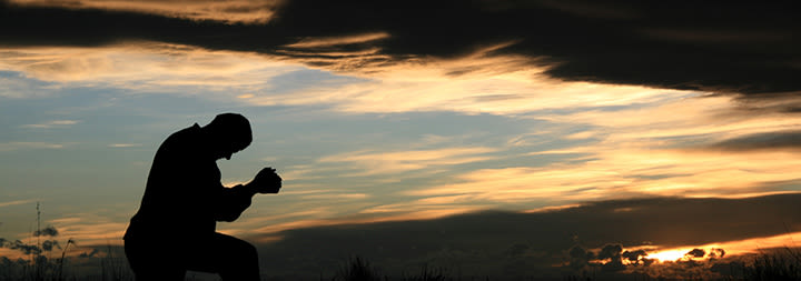 A man kneels in prayer against a dramatic sunset backdrop.