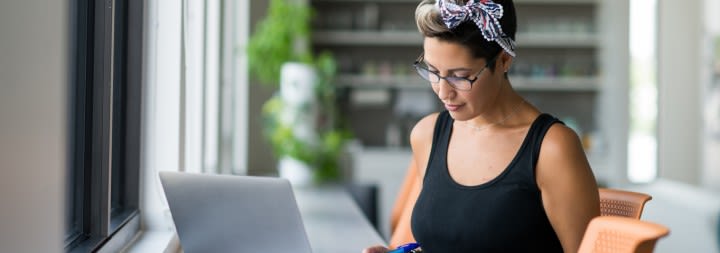 Woman works on a laptop near a sunlit window.