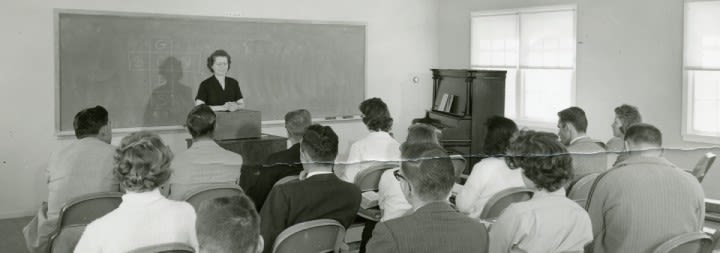 An old b/w photograph of a teacher at the front of a class of adult learners.