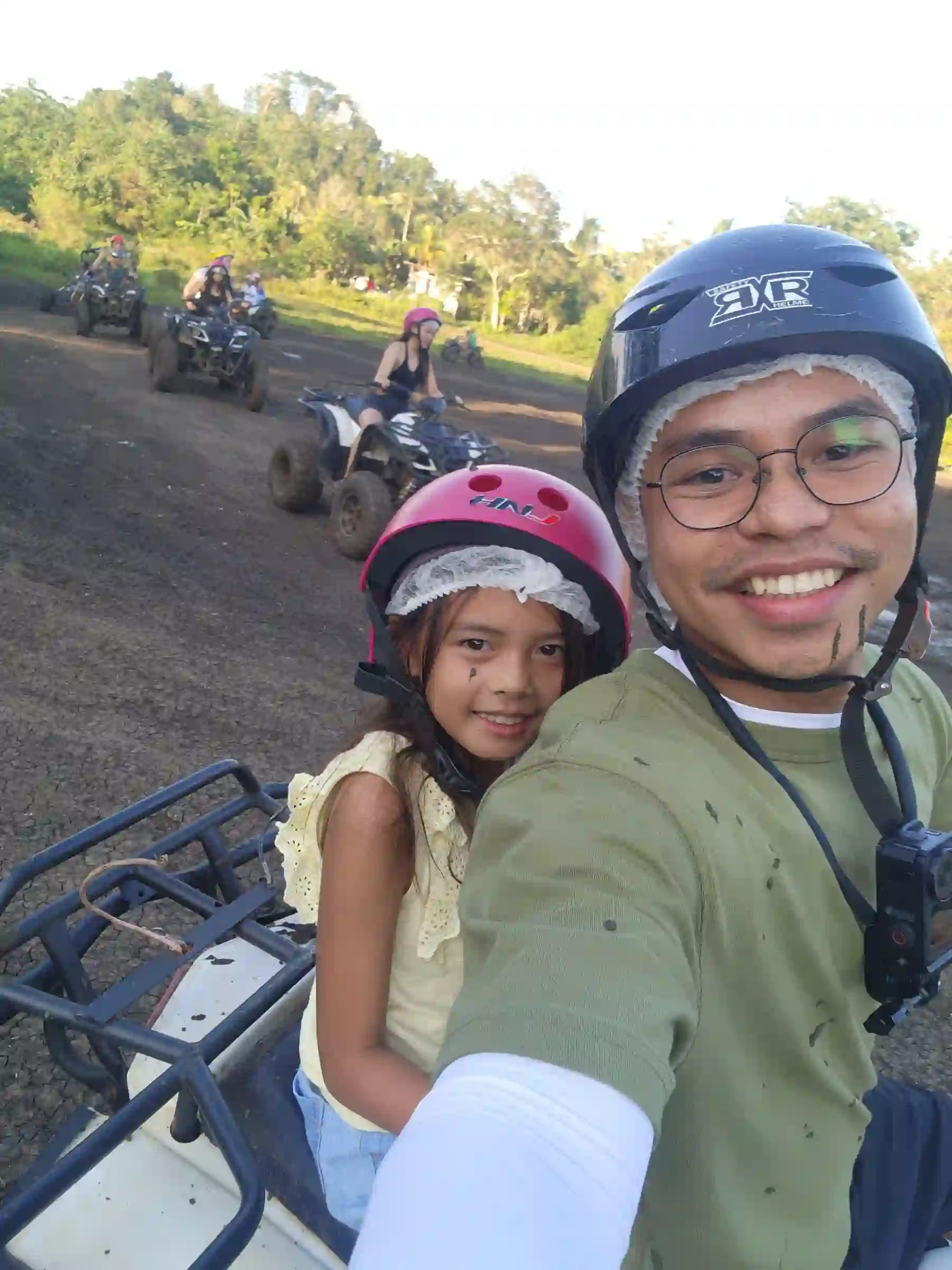 Nelmar Serondo Atanoza in black helmet and green shirt taking a selfie with a young girl in a pink helmet while riding an ATV on a dirt road near the Chocolate Hills in Carmen Bohol