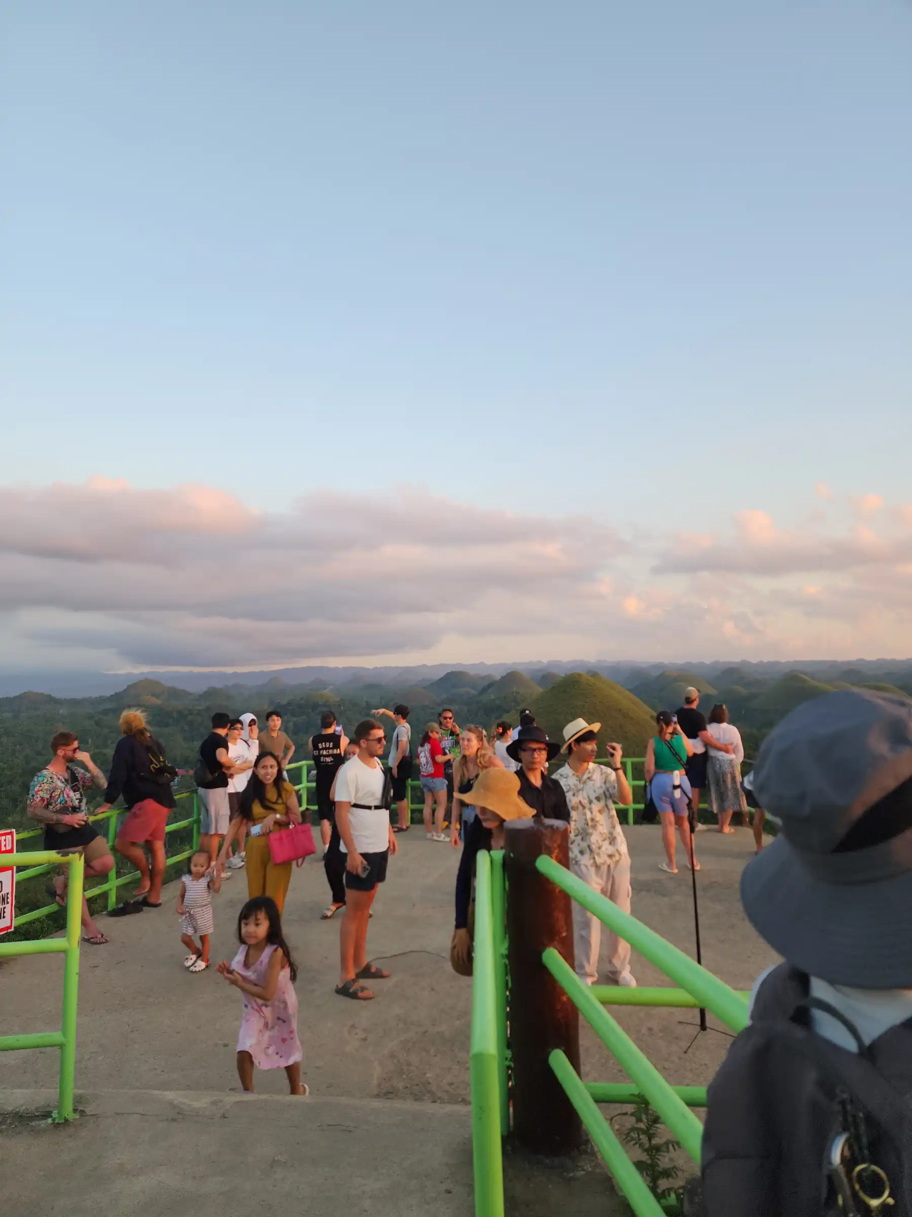 Tourists standing along the green railing of the Chocolate Hills Complex observation deck at sunset with dozens of green conical hills visible in the background at Carmen Bohol