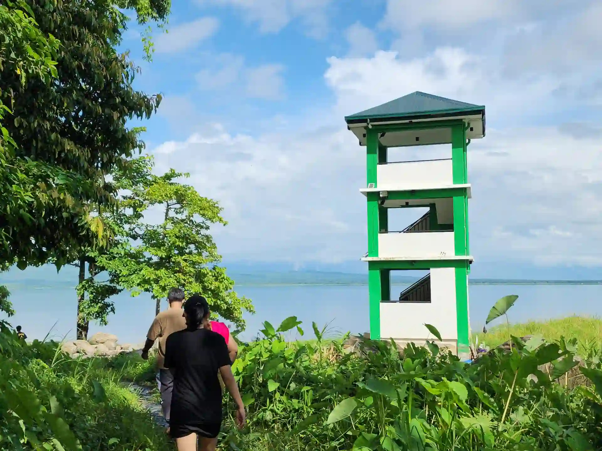 Three visitors walking along a grassy lakeside path toward a white and green observation tower at Naujan Lake in Mabuhay II Socorro Oriental Mindoro
