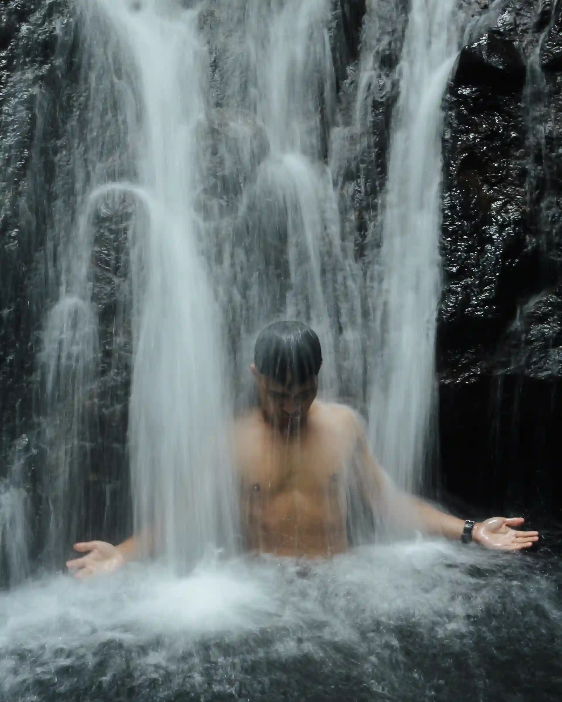 A man sitting under the cascading streams of Magkayad Falls with both arms extended and palms open in the pool at Barangay Maputi San Isidro Davao Oriental