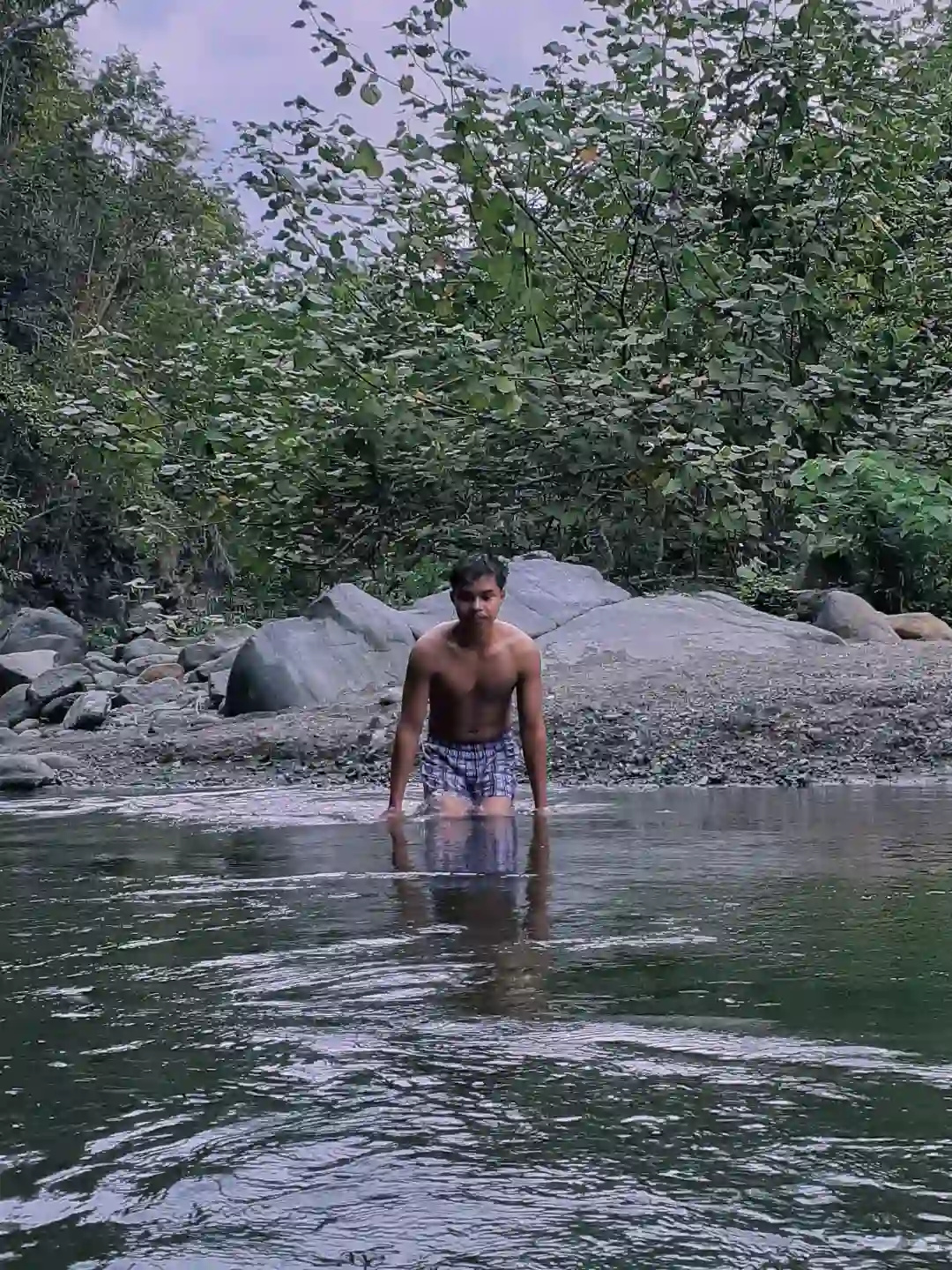 A young man wading knee deep in a clear rocky river with large boulders and green trees on the bank in Barangay Maputi San Isidro Davao Oriental