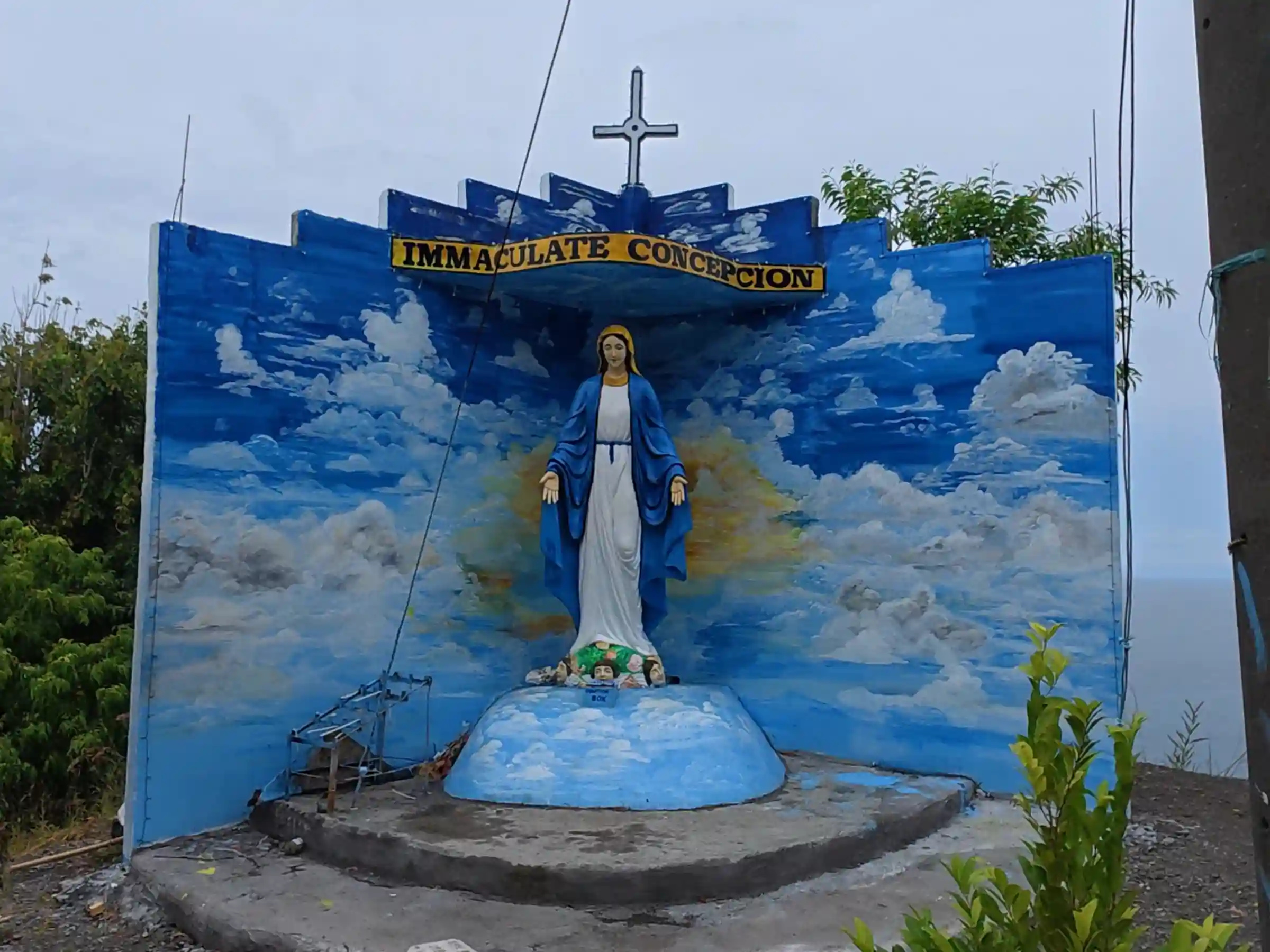 Painted concrete shrine of the Immaculate Concepcion with a hand painted sky backdrop and metal cross at the Lanca Cabuaya Road overlooking point in Mati Davao Oriental