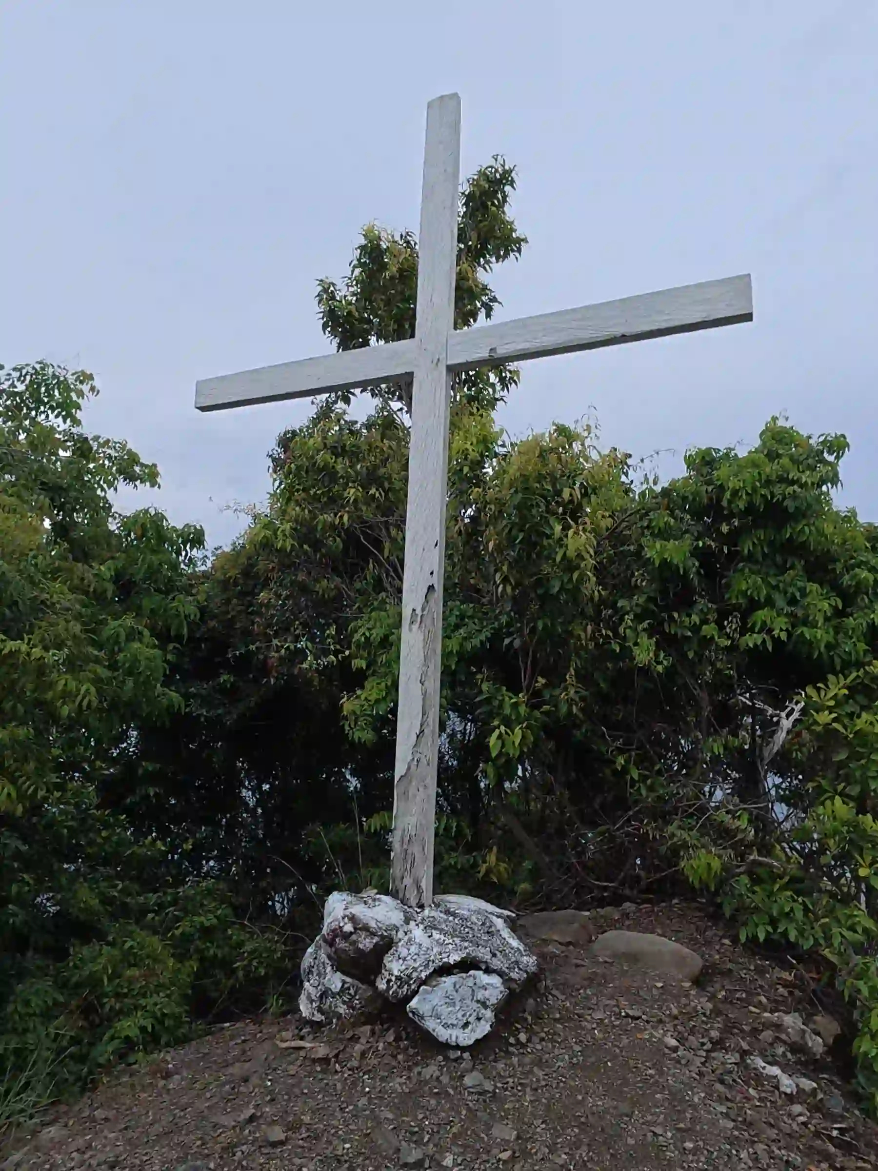 White painted wooden cross planted in whitewashed rocks on a hillside above the Lanca Cabuaya Road overlooking point in Mati Davao Oriental with trees behind