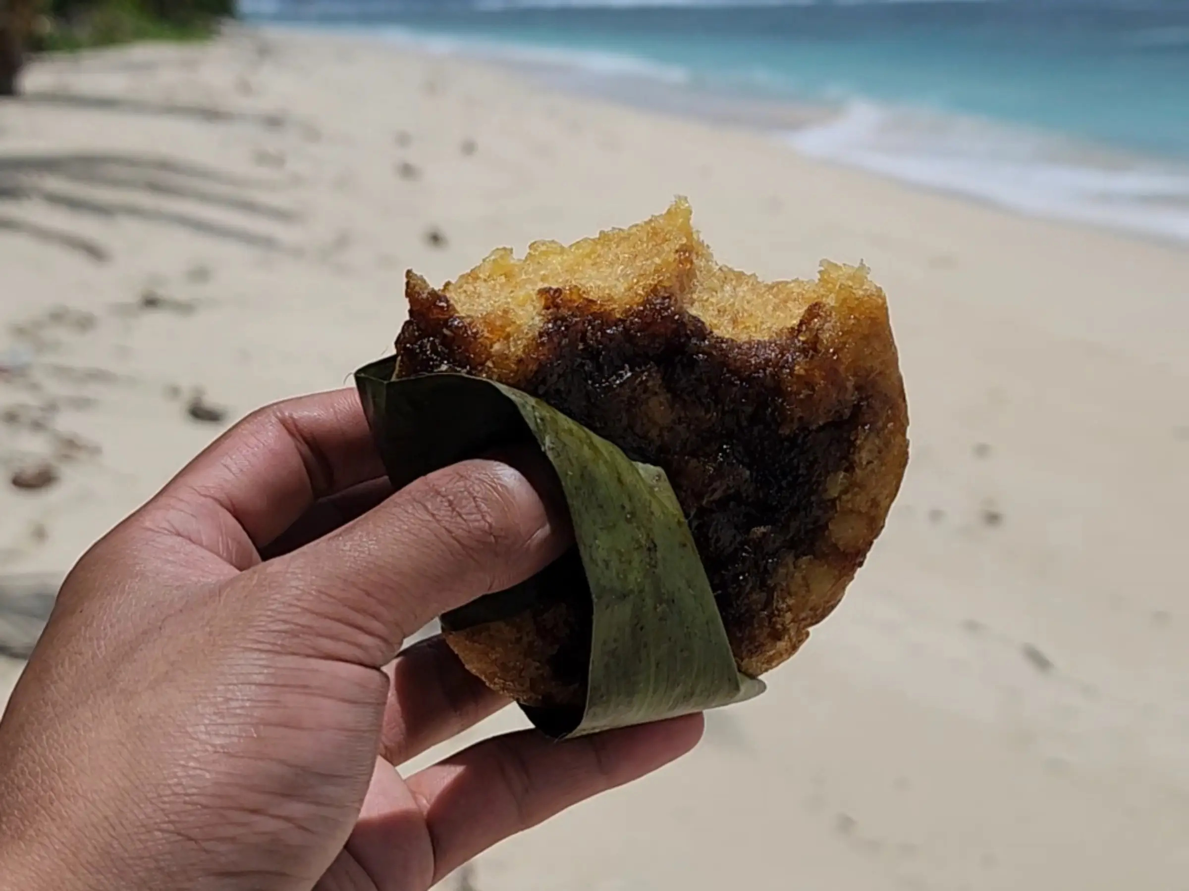 A hand holding a piece of puto cassava wrapped in banana leaf with Lanca Beach and a forested headland visible in the background