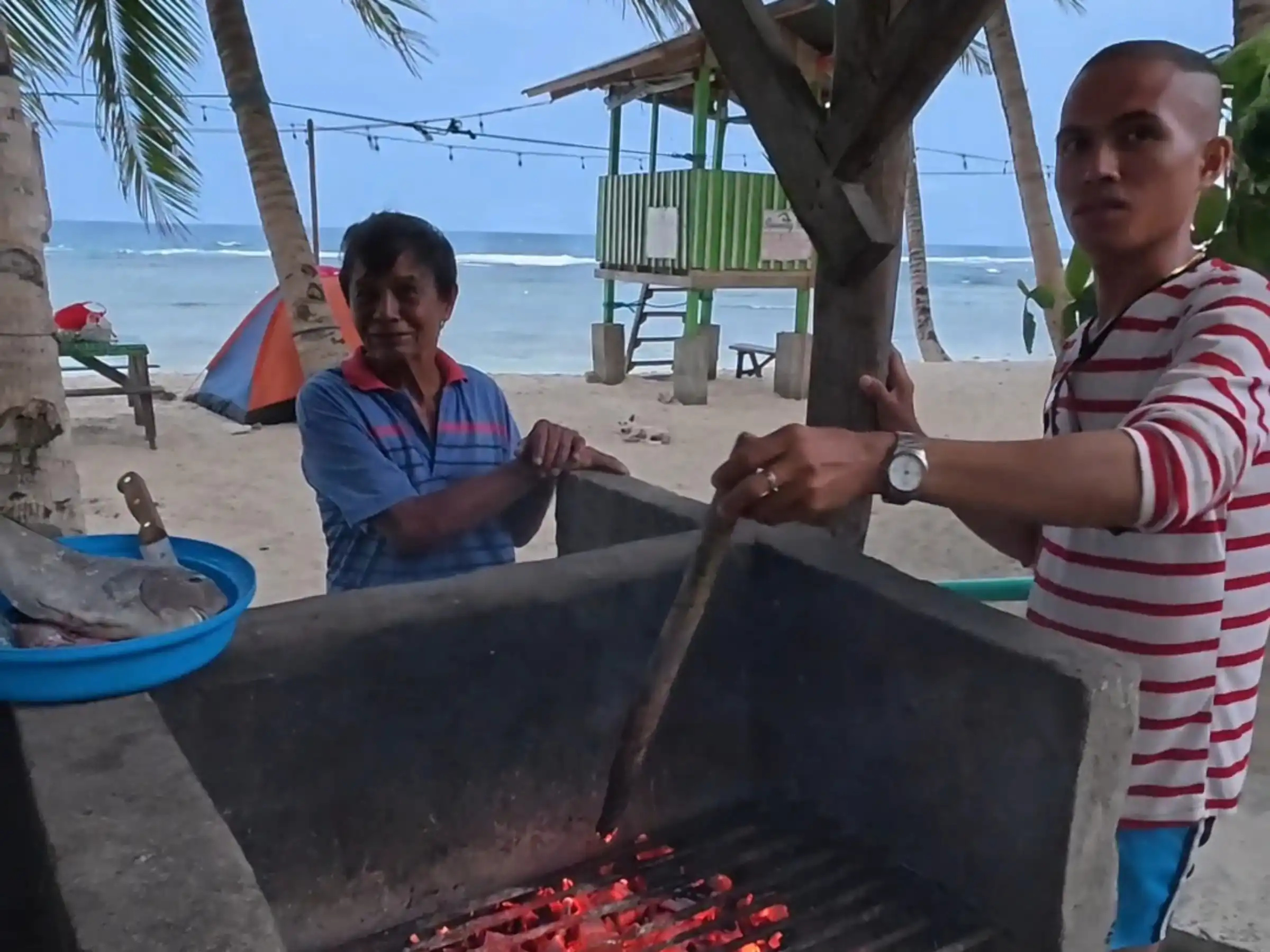 Two men tending a concrete charcoal grill with glowing coals under a corrugated roof near Lanca beach in Mati Davao Oriental