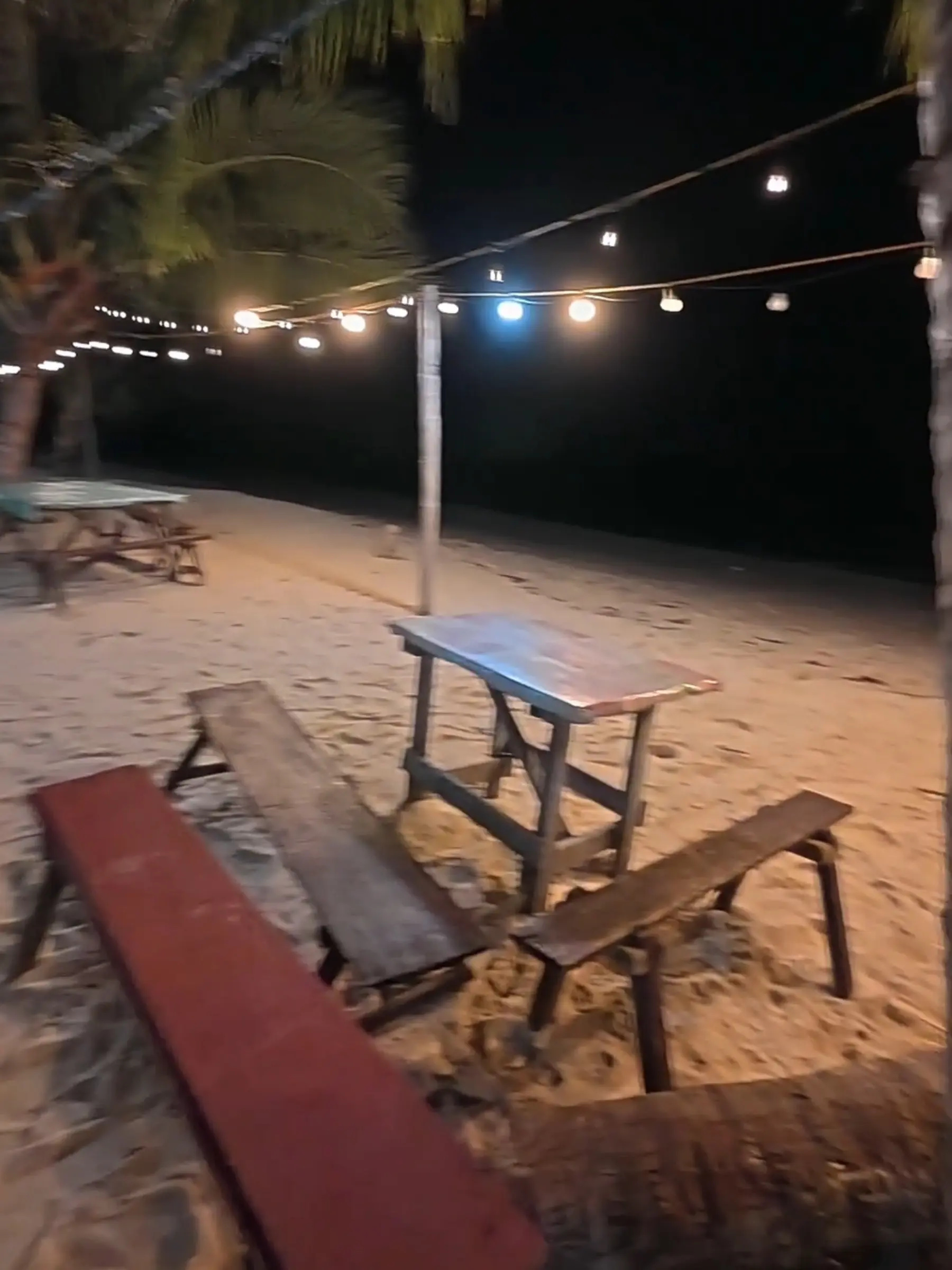 Wooden picnic tables on pale sand at night under string lights at Lanca Shoreline Camping Site in Mati Davao Oriental