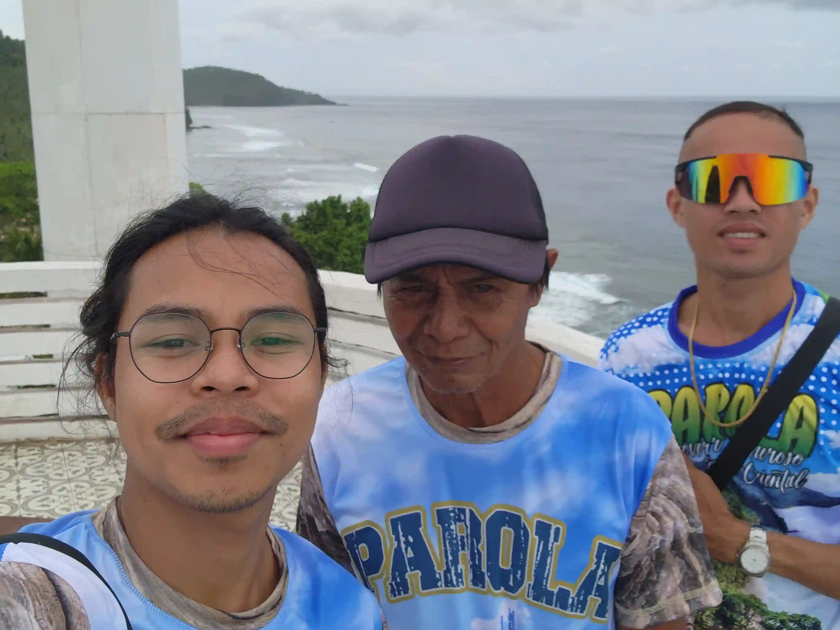 Three men wearing matching Cape San Agustin Parola souvenir shirts taking a selfie at the lighthouse viewing deck in Governor Generoso Davao Oriental with the Pacific coastline and a distant headland visible behind them