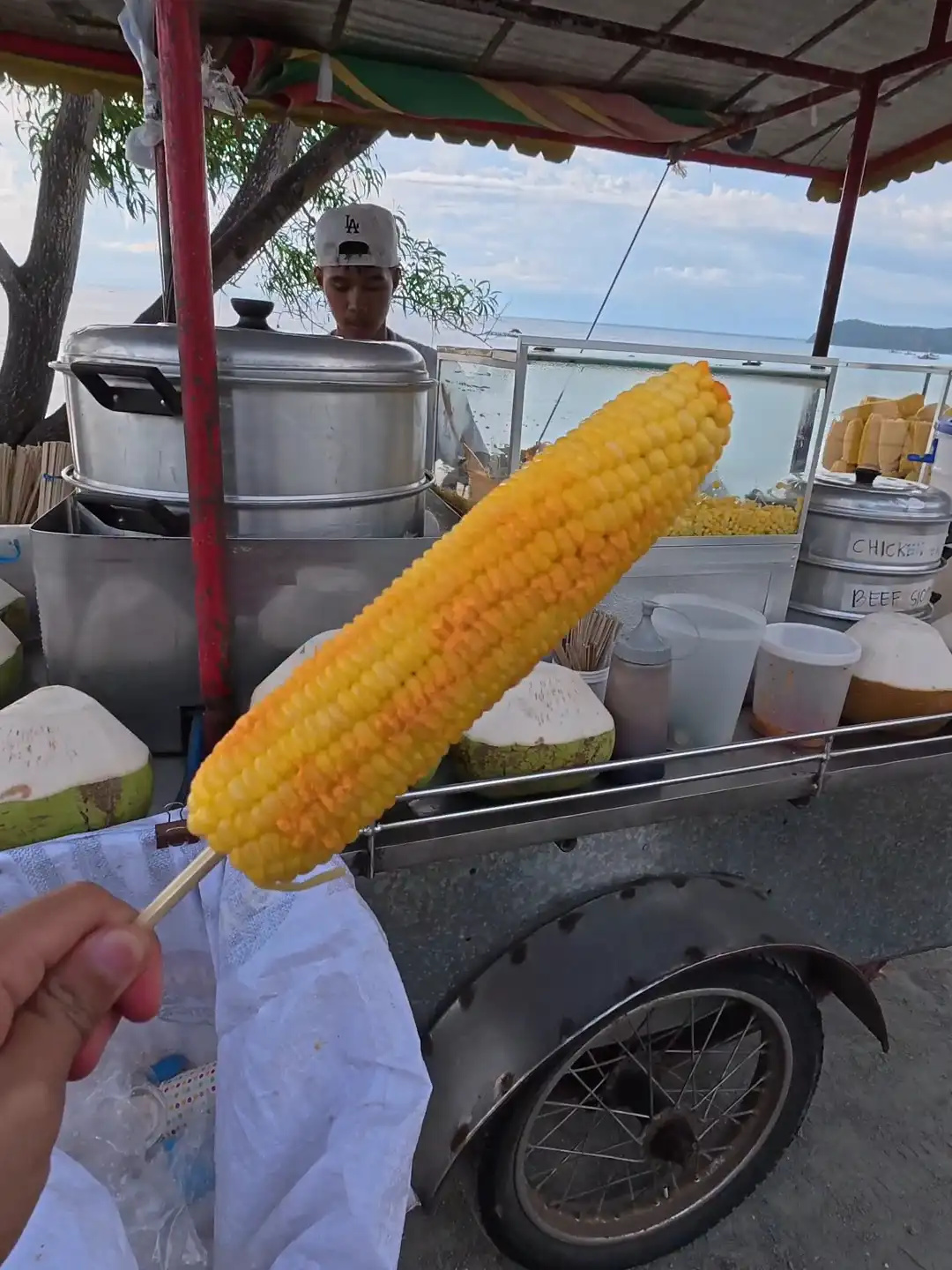 A hand holding a boiled young corn on a stick in front of a roadside tricycle food vendor cart at Punta Abijod in San Isidro Davao Oriental