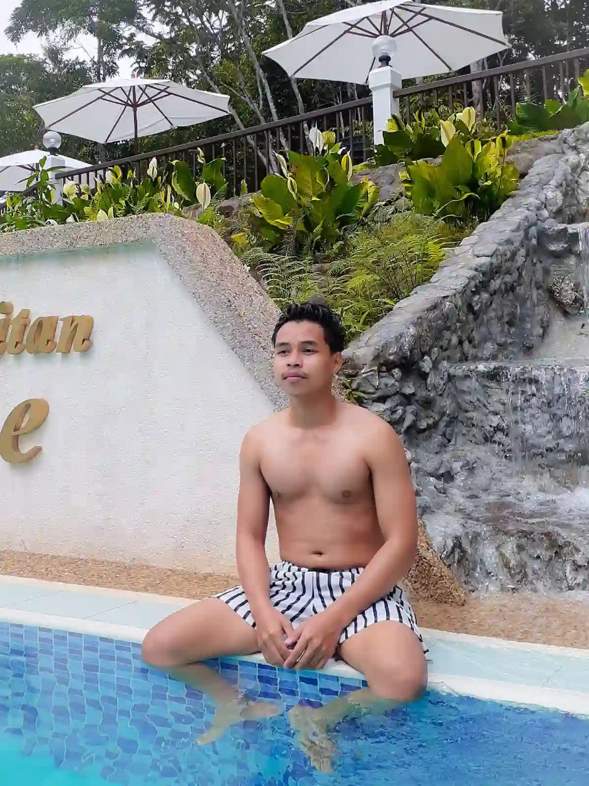 Man sitting at the pool ledge directly beside the Mt. Hamiguitan Escape stone marker with a working waterfall feature to the right and white umbrellas and dense forest trees behind in San Isidro Davao Oriental