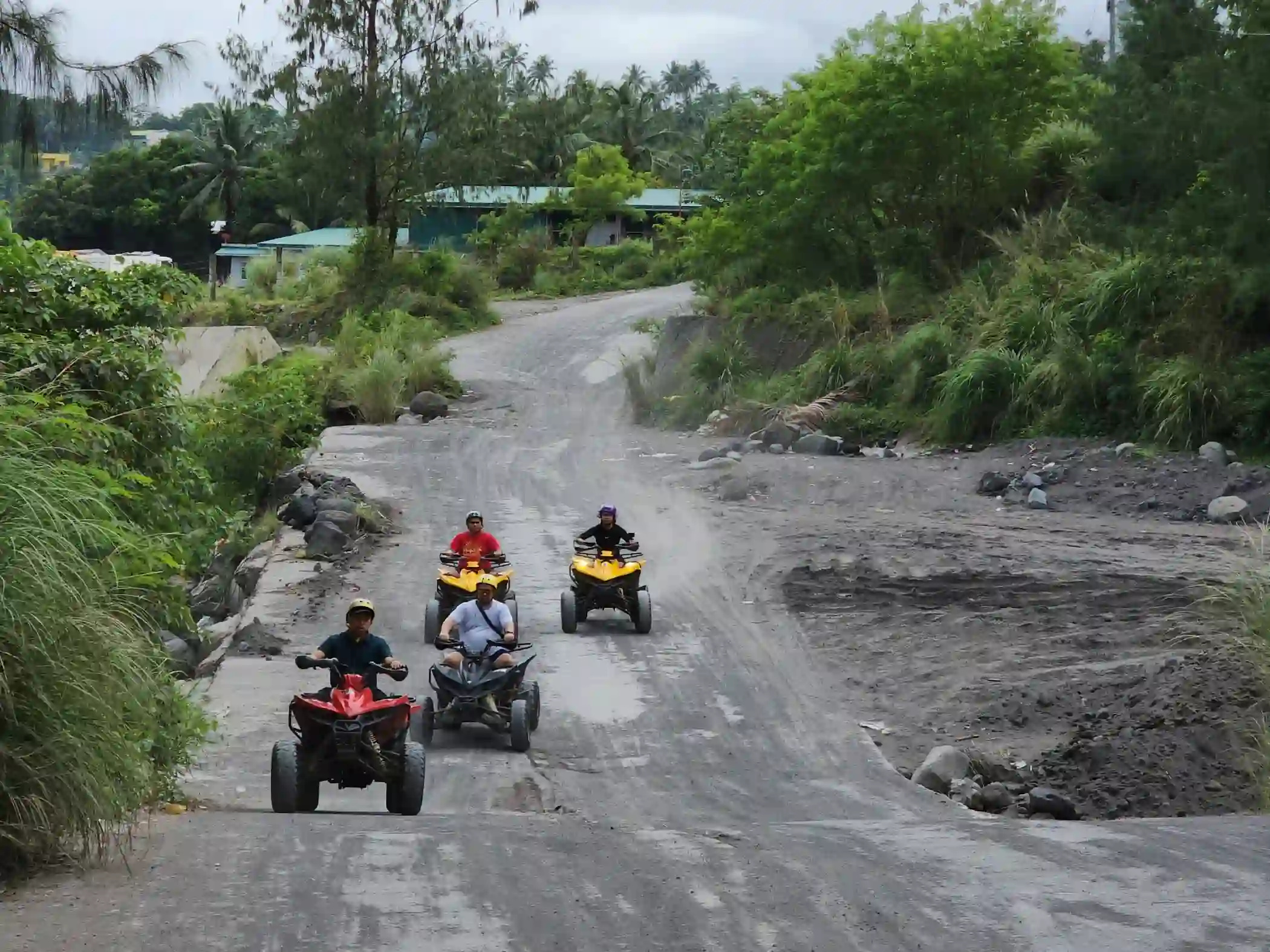 Four ATV riders on red and yellow ATVs riding along a wide volcanic gravel path flanked by trees and shrubs on the route to Green Lava in Daraga Albay