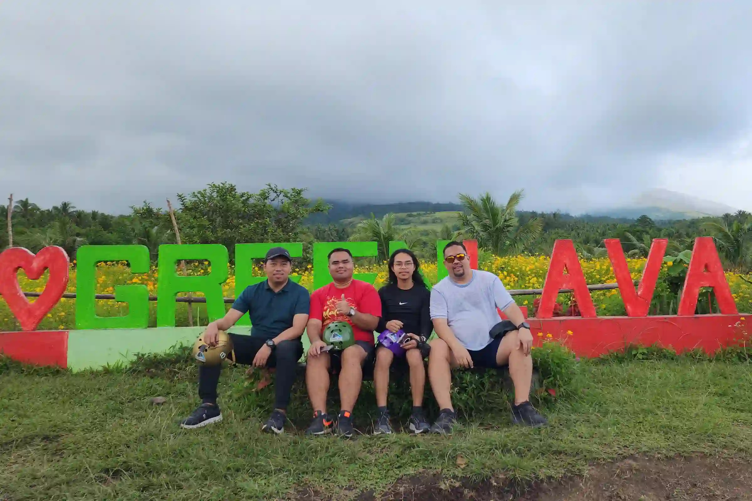 Four riders sitting on the Green Lava painted sign with yellow flowers and the cloud covered peak of Mayon Volcano visible in the background in Daraga Albay