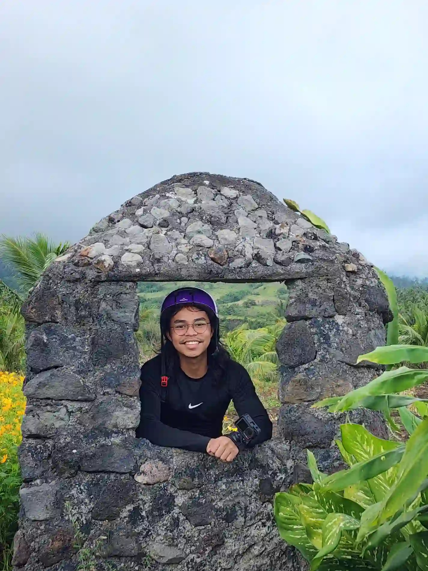 Nelmar Atanoza in a purple helmet leaning through the window of a small dark volcanic stone arch structure with green hills and clouds visible behind him at Green Lava in Daraga Albay