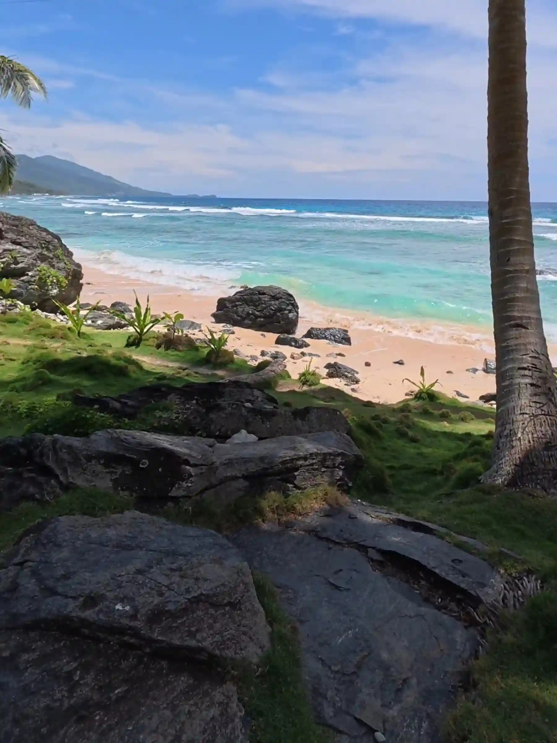 Coconut trunk in foreground with layered black rock formations and bermuda grass leading to creamy orange sand and turquoise ocean at Payag ni Jose in Lanca Mati Davao Oriental