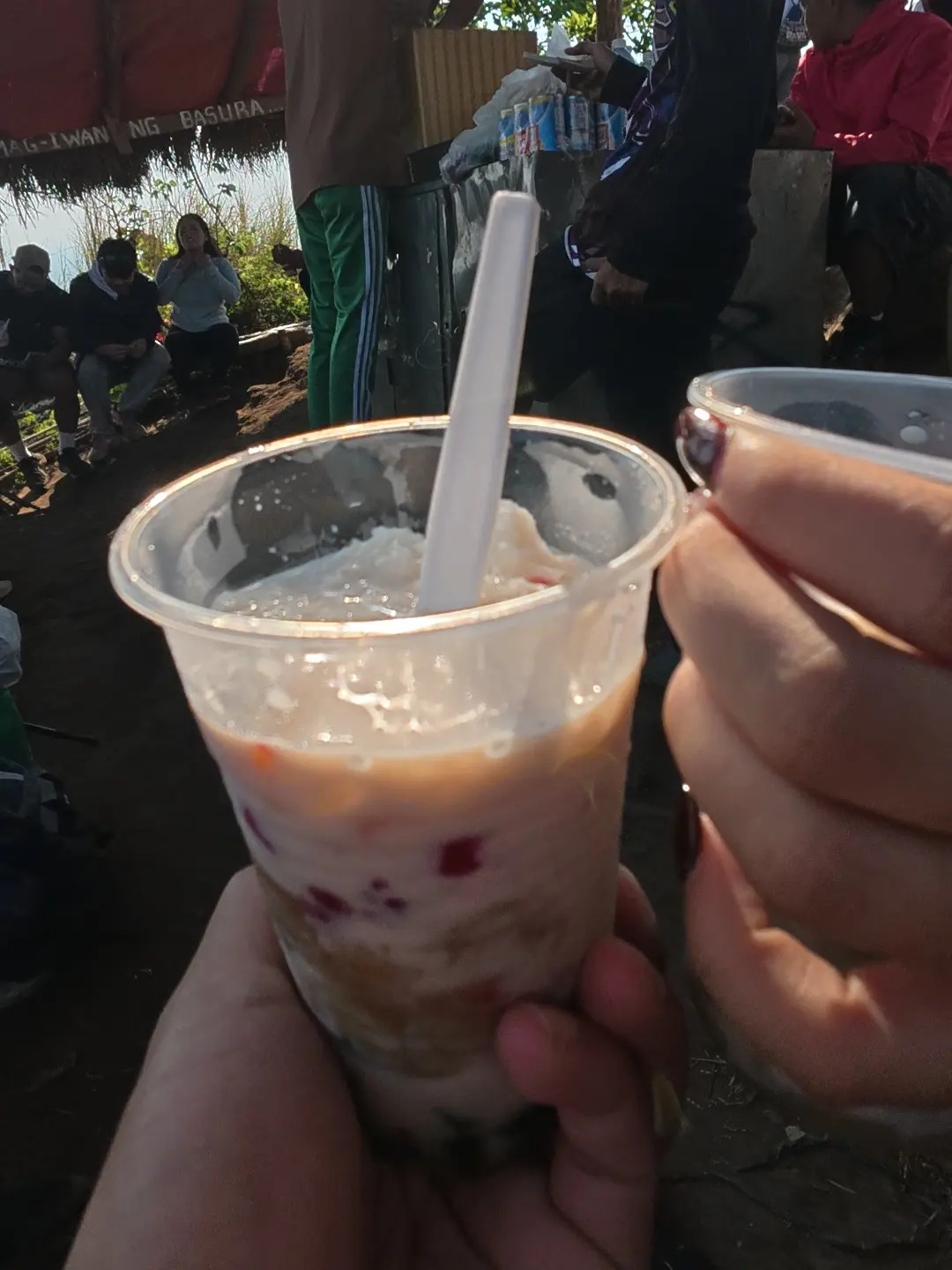 Two hands holding plastic cups of halo-halo at the summit food stall of Mt. Talamitam with other hikers and vendors visible in the background in Nasugbo Batangas