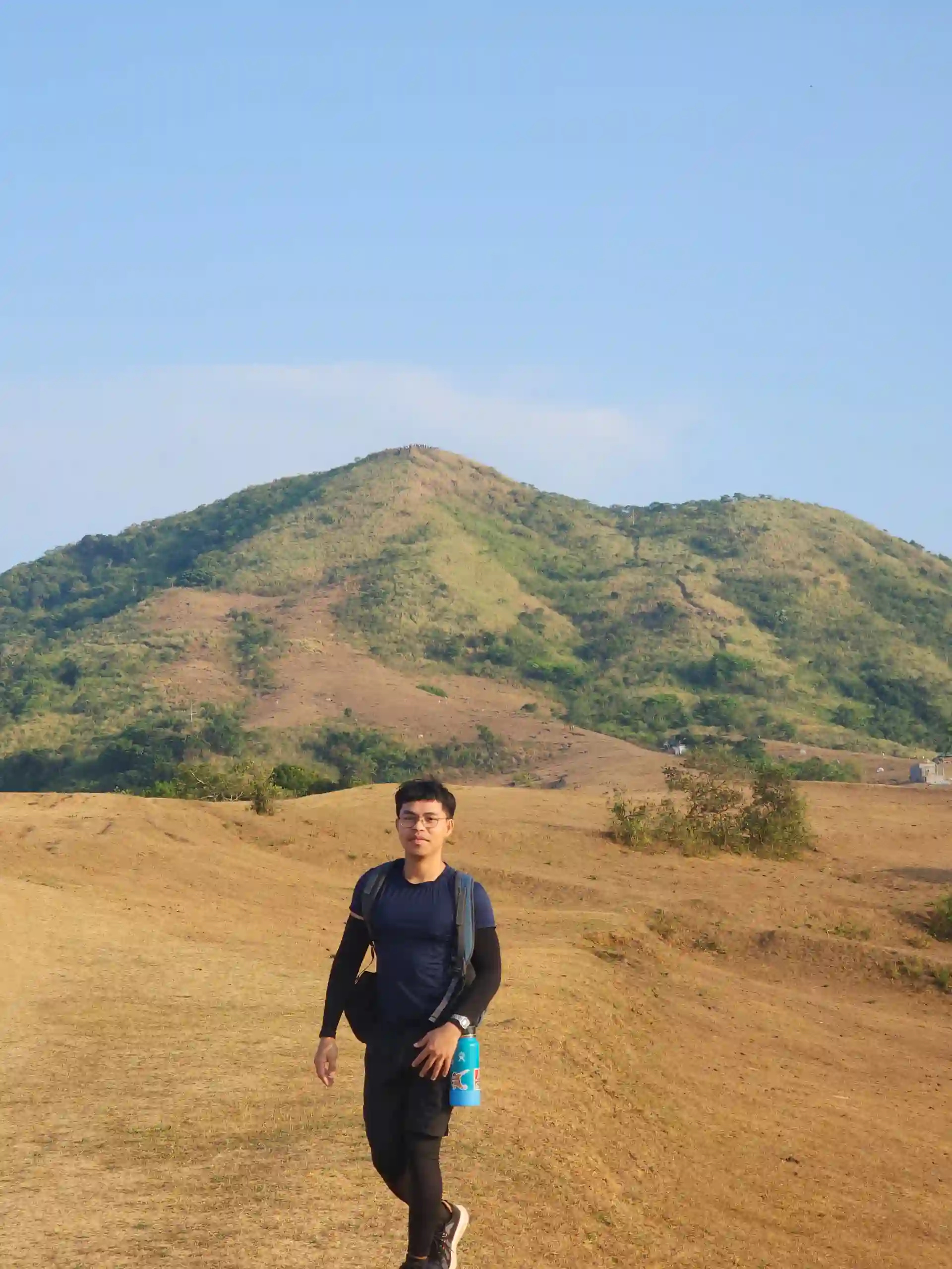 A lone hiker with a blue backpack and water bottle standing on an open ridge looking toward brown dry grass hills under clear blue sky at Mt. Talamitam Nasugbo Batangas