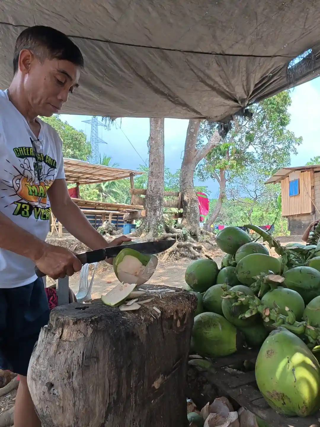 A male vendor chopping open a green young coconut with a large knife on a wooden stump under a tarpaulin shade at the Mt. Talamitam jump-off area in Nasugbo Batangas