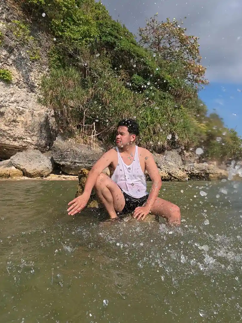 Carlo Albento in a white tank top sitting on a submerged rock at Talim Beach in Lian Batangas as a wave hits and sends water splashing around him with the limestone cliff in the background