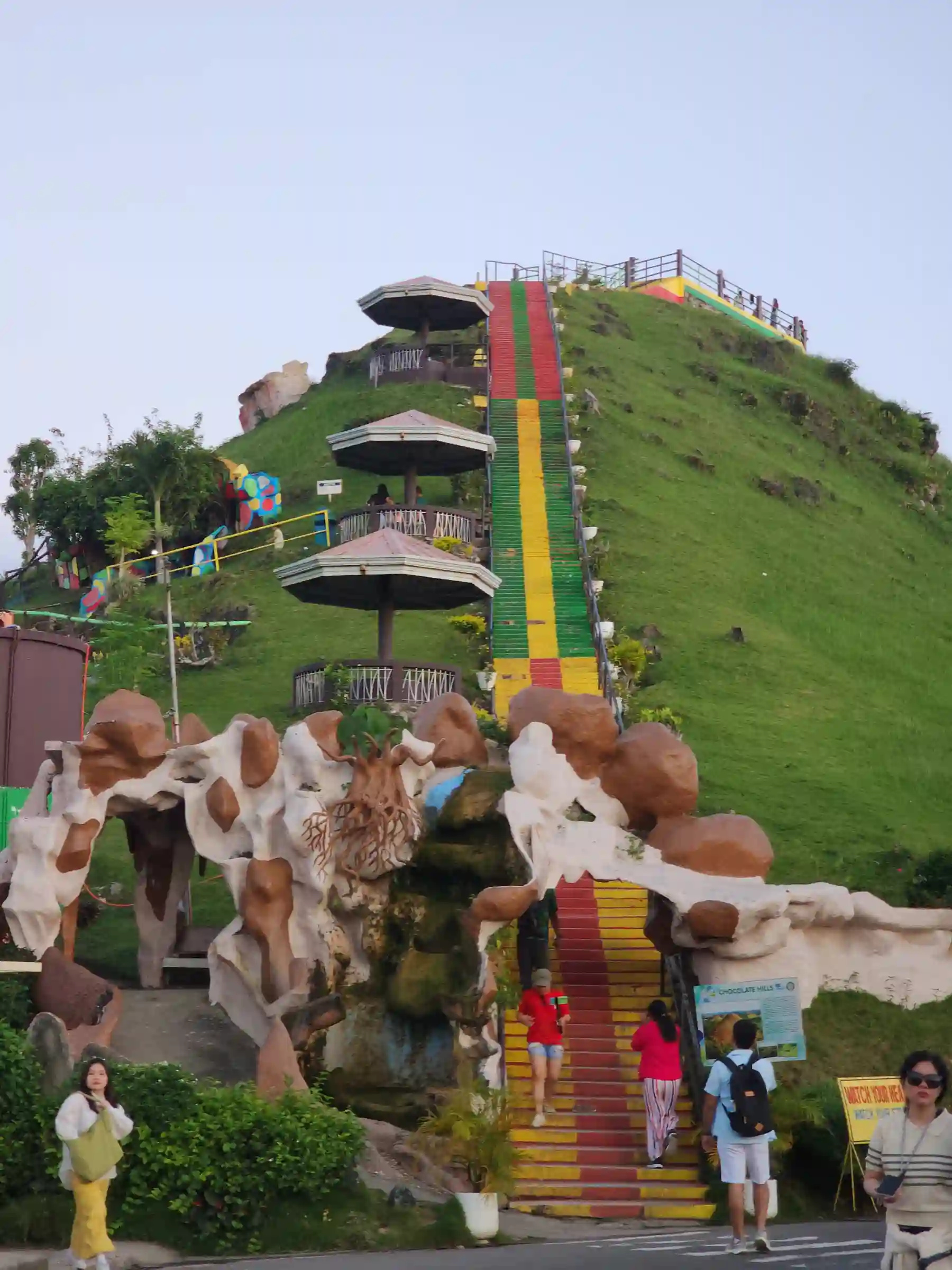 Colorful red yellow and green stairs leading up a grassy Chocolate Hills viewpoint complex in Carmen Bohol with concrete sculptures at the base and hexagonal pavilions along the climb