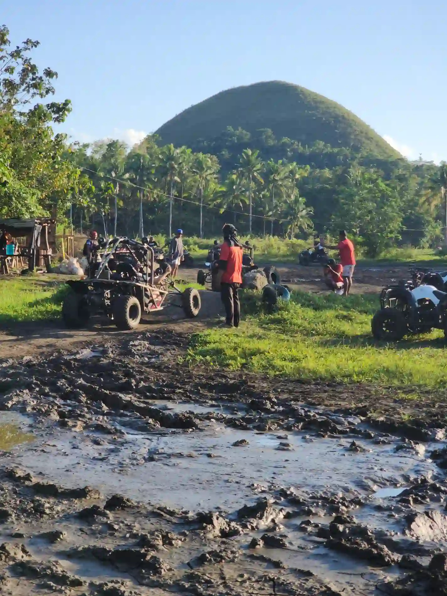 ATV buggies and quad bikes parked at a muddy staging area at the base of the Chocolate Hills in Carmen Bohol with a green conical hill rising behind operators in orange shirts