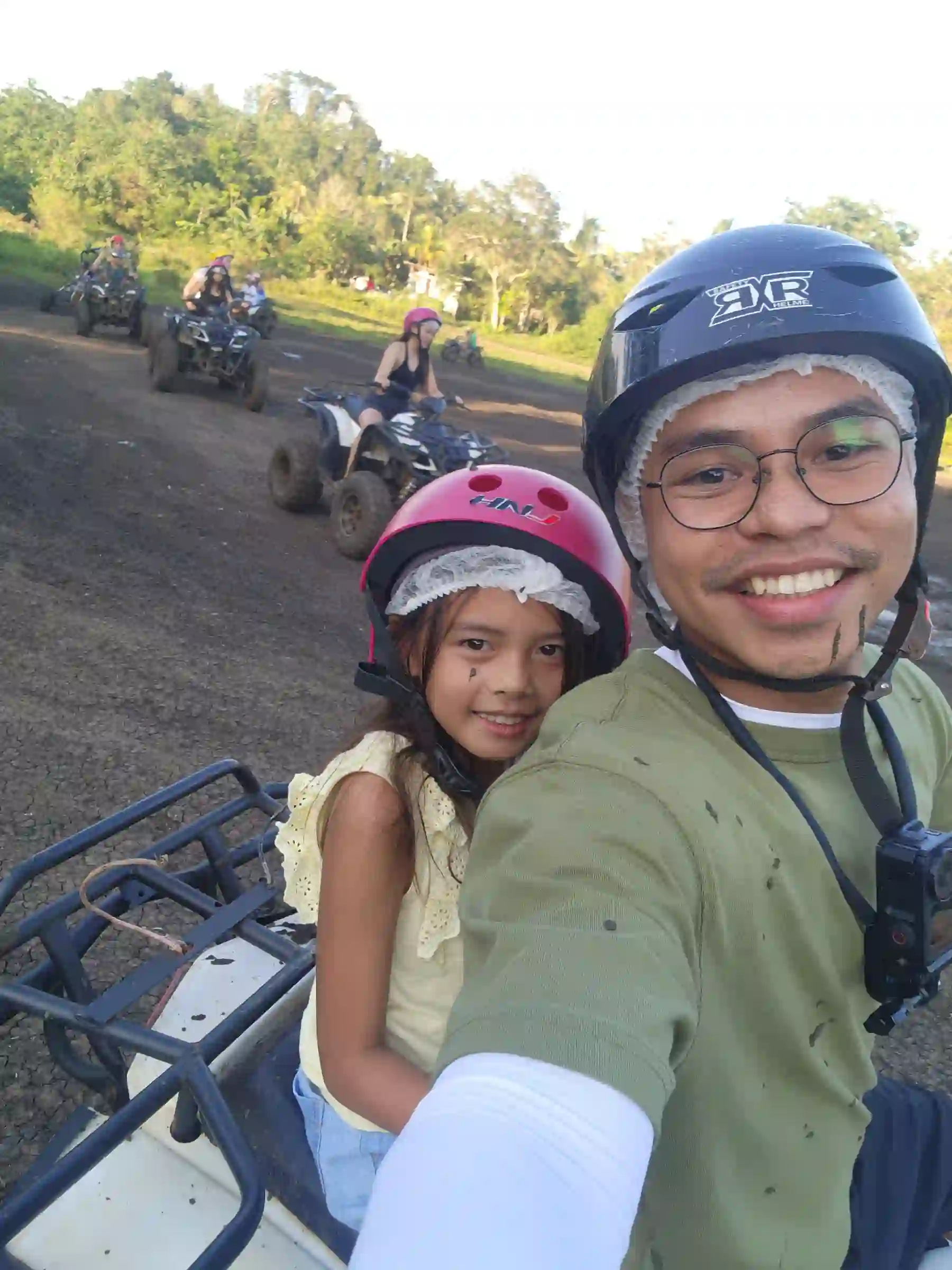 Nelmar Serondo Atanoza in black helmet and green shirt taking a selfie with a young girl in a pink helmet while riding an ATV on a dirt road near the Chocolate Hills in Carmen Bohol