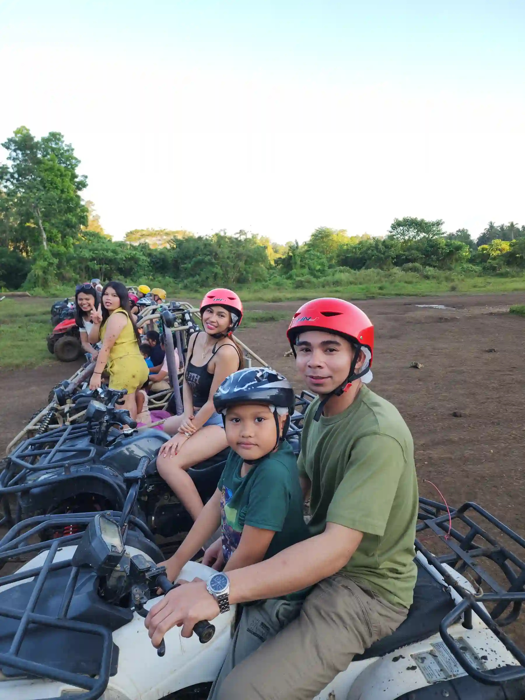 Group of adults and a child in helmets sitting on ATV units lined up at the staging area with green trees behind them near the Chocolate Hills Carmen Bohol