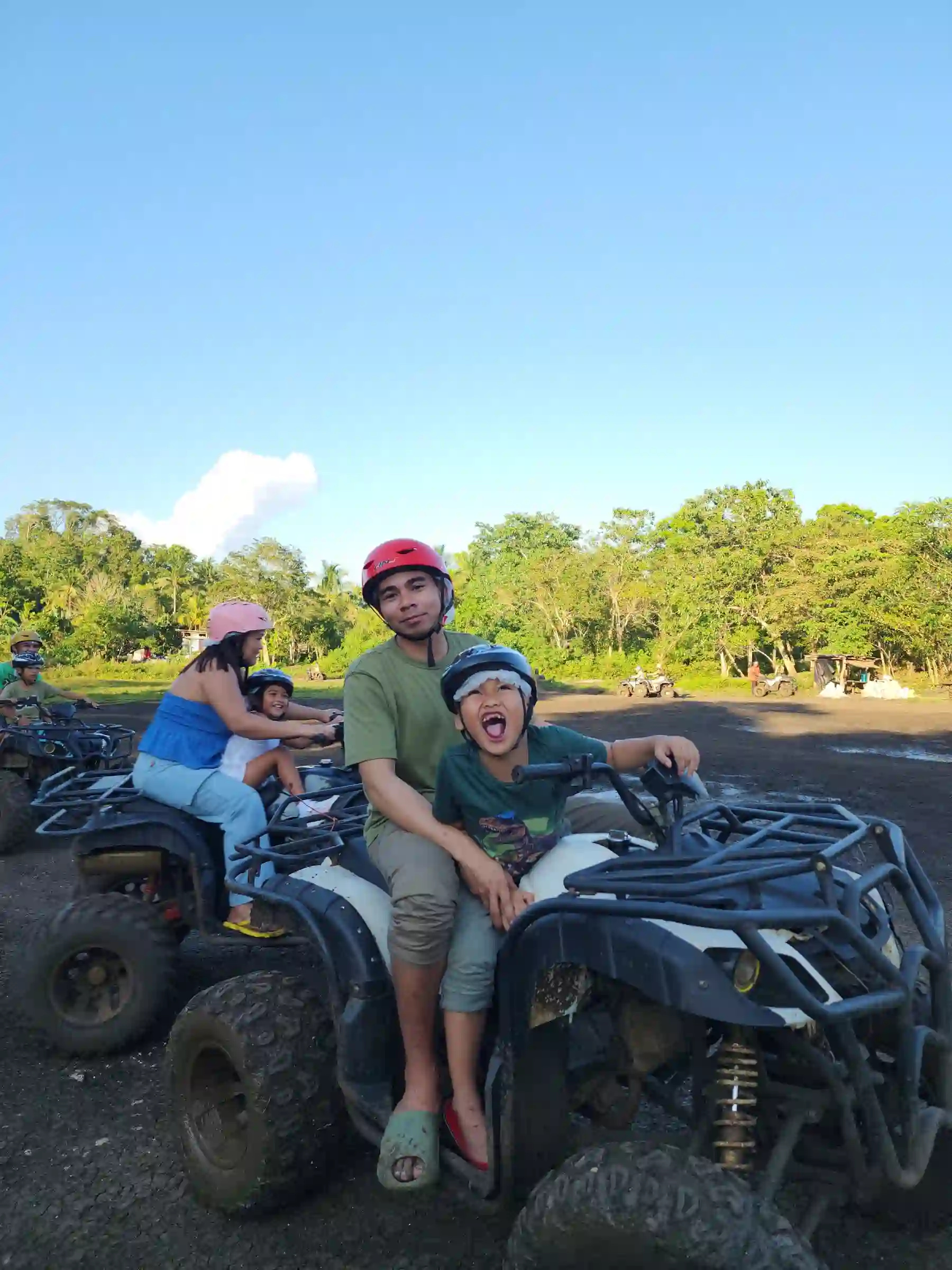 Marby Serondo Atanoza in red helmet and green shirt sitting on an ATV with a laughing child in a blue helmet against a clear blue sky at the Chocolate Hills ATV area in Carmen Bohol