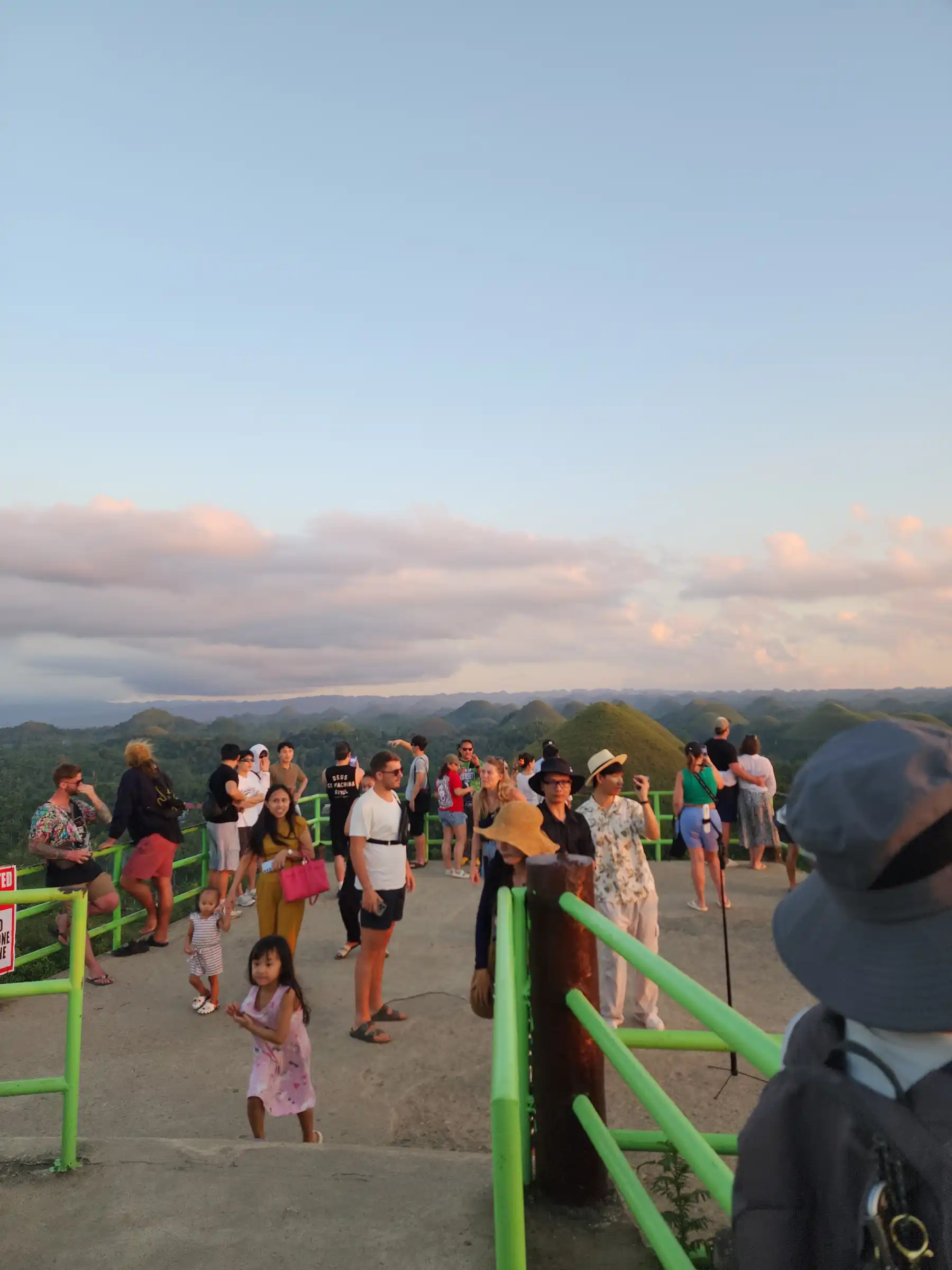 Tourists standing along the green railing of the Chocolate Hills Complex observation deck at sunset with dozens of green conical hills visible in the background at Carmen Bohol