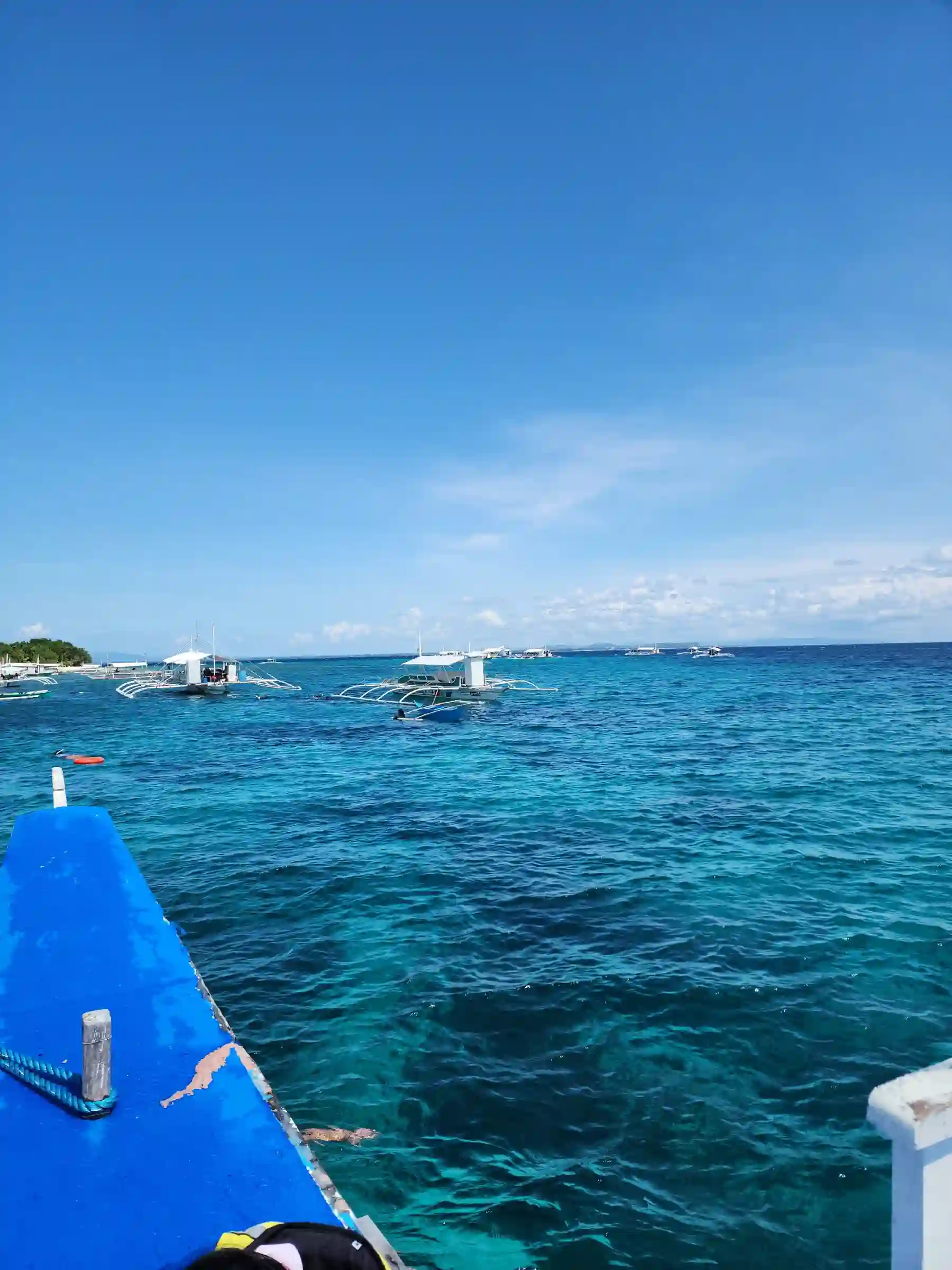 View from the bow of a bangka approaching Balicasag Island Bohol showing outrigger poles framing the green forested island and turquoise water