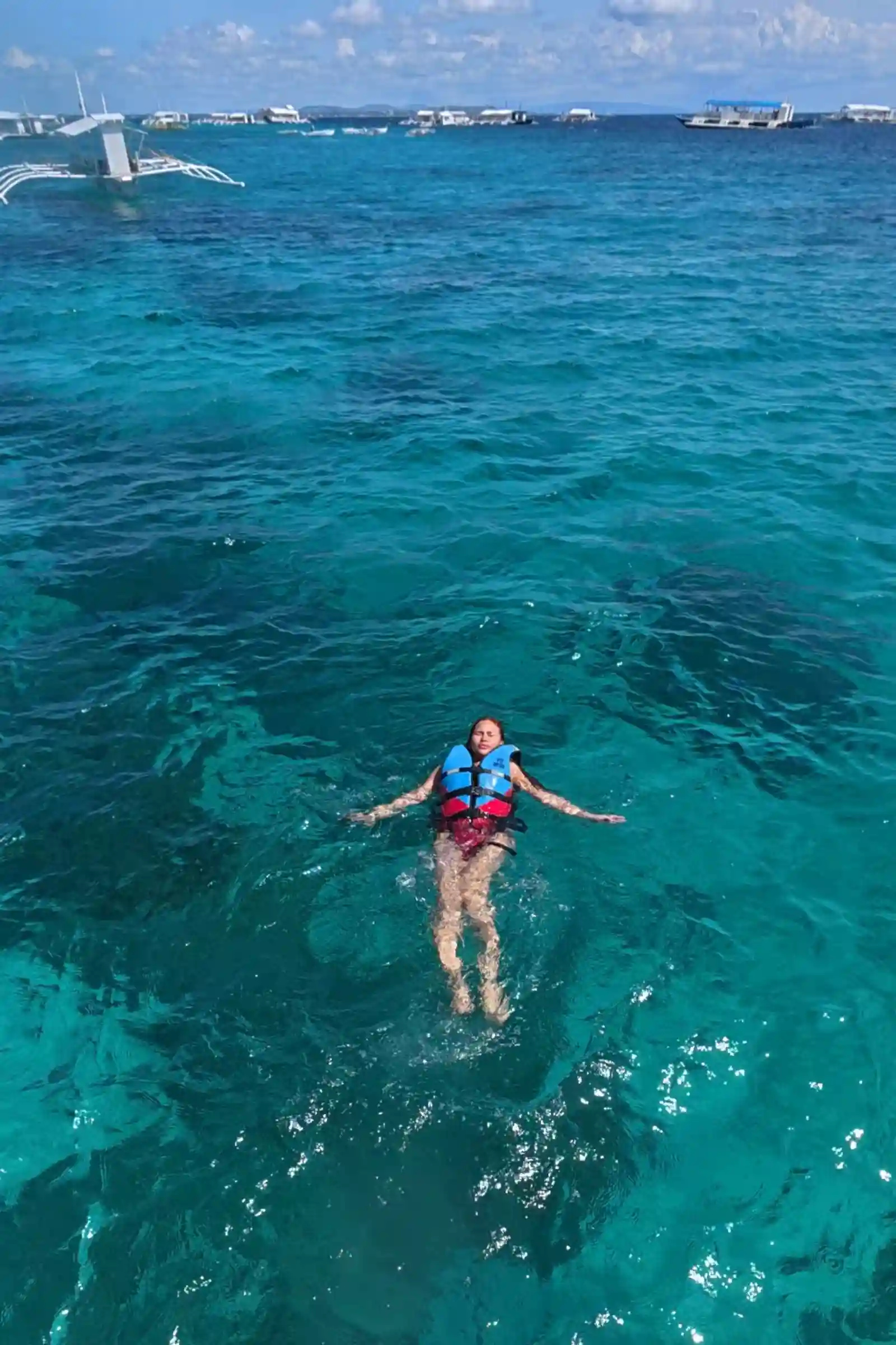Snorkeler Angelie Inangkilat in blue and red life vest floating on her back in turquoise water near Balicasag Island Bohol with bangkas in the background