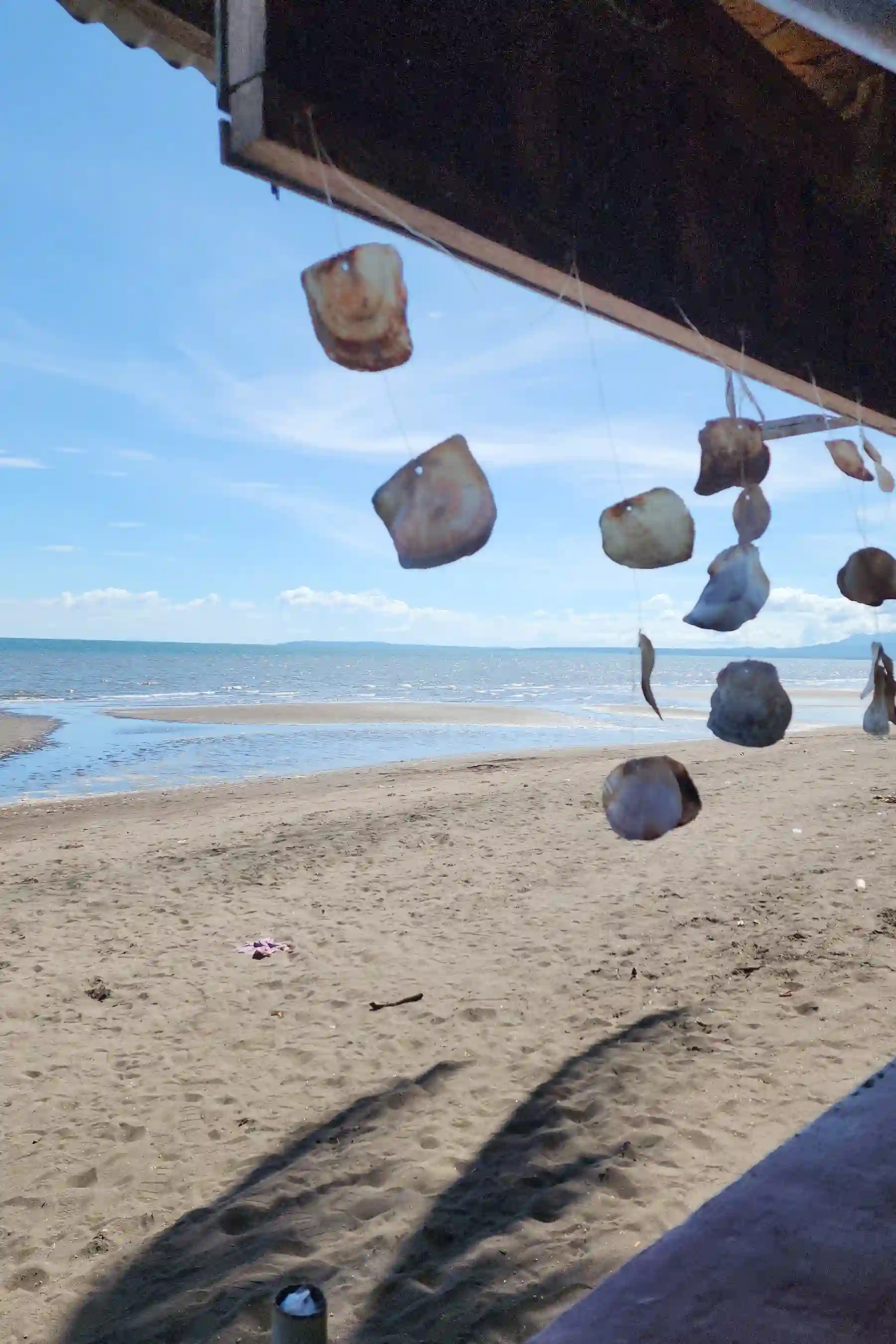 Shell wind chime hanging from the eave of a nipa hut with dark sand beach and sea visible in the background at Villa Del Prado Sariaya Quezon