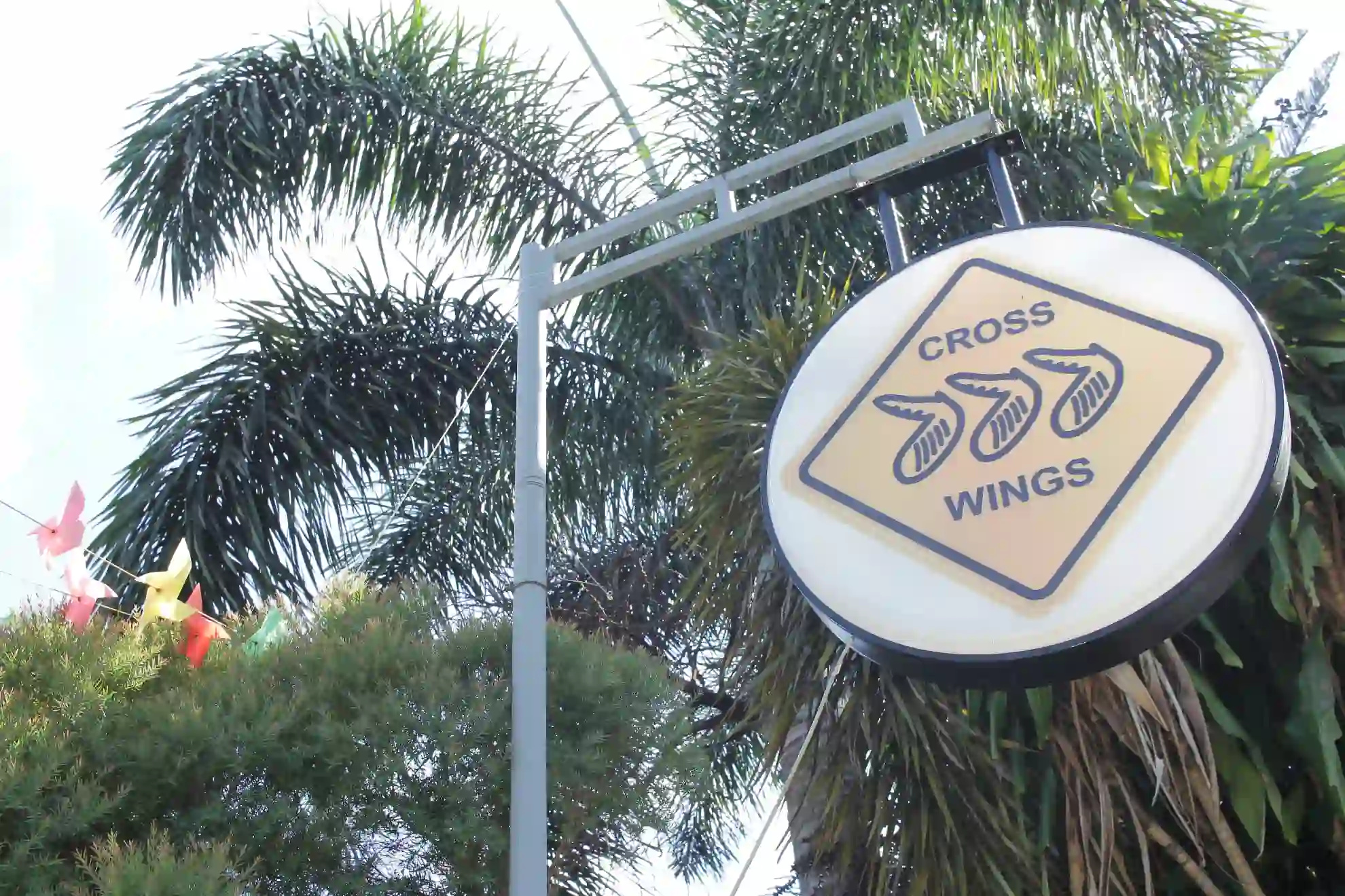 Low angle view of the Crosswings round hanging sign against a blue sky with colorful pinwheels and trees in Alfonso Cavite