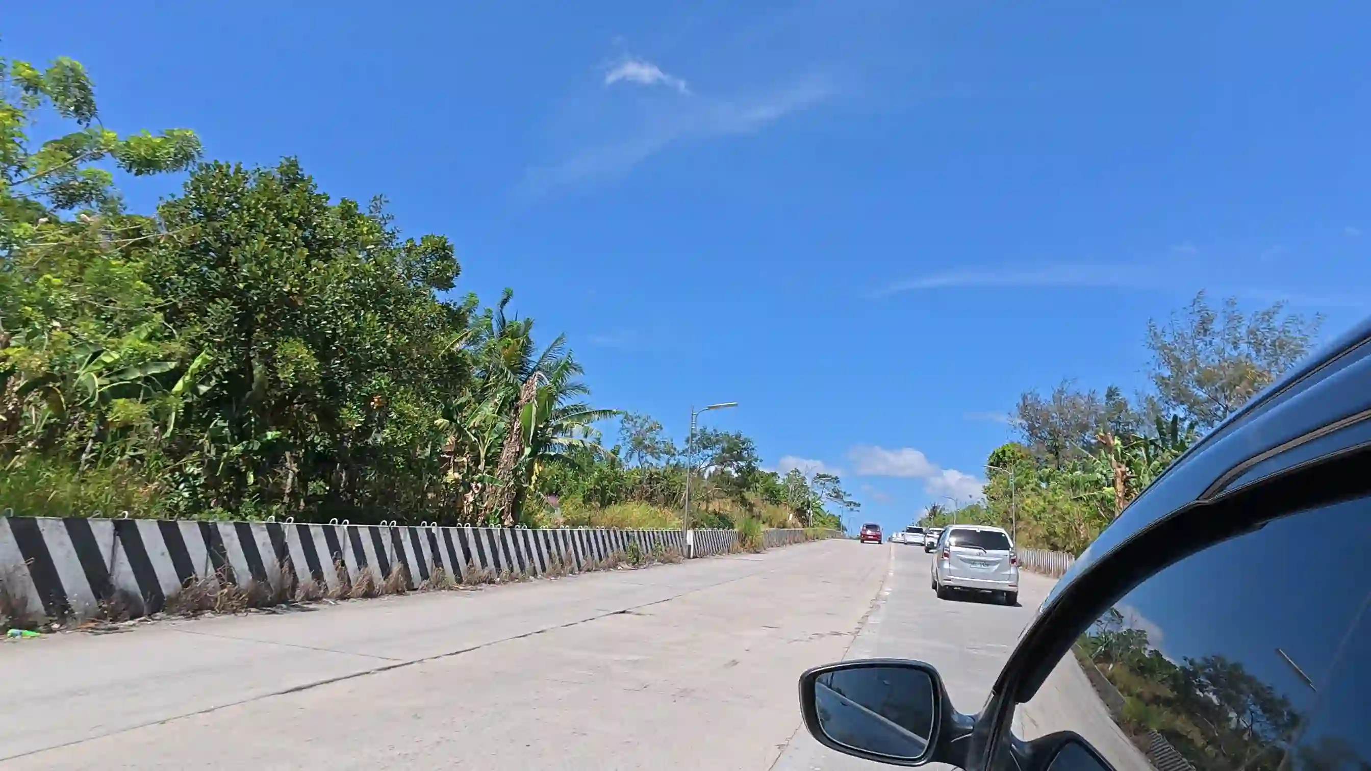 View from inside a car driving along the Tagaytay bypass road lined with tropical trees and a striped barrier wall under a blue sky