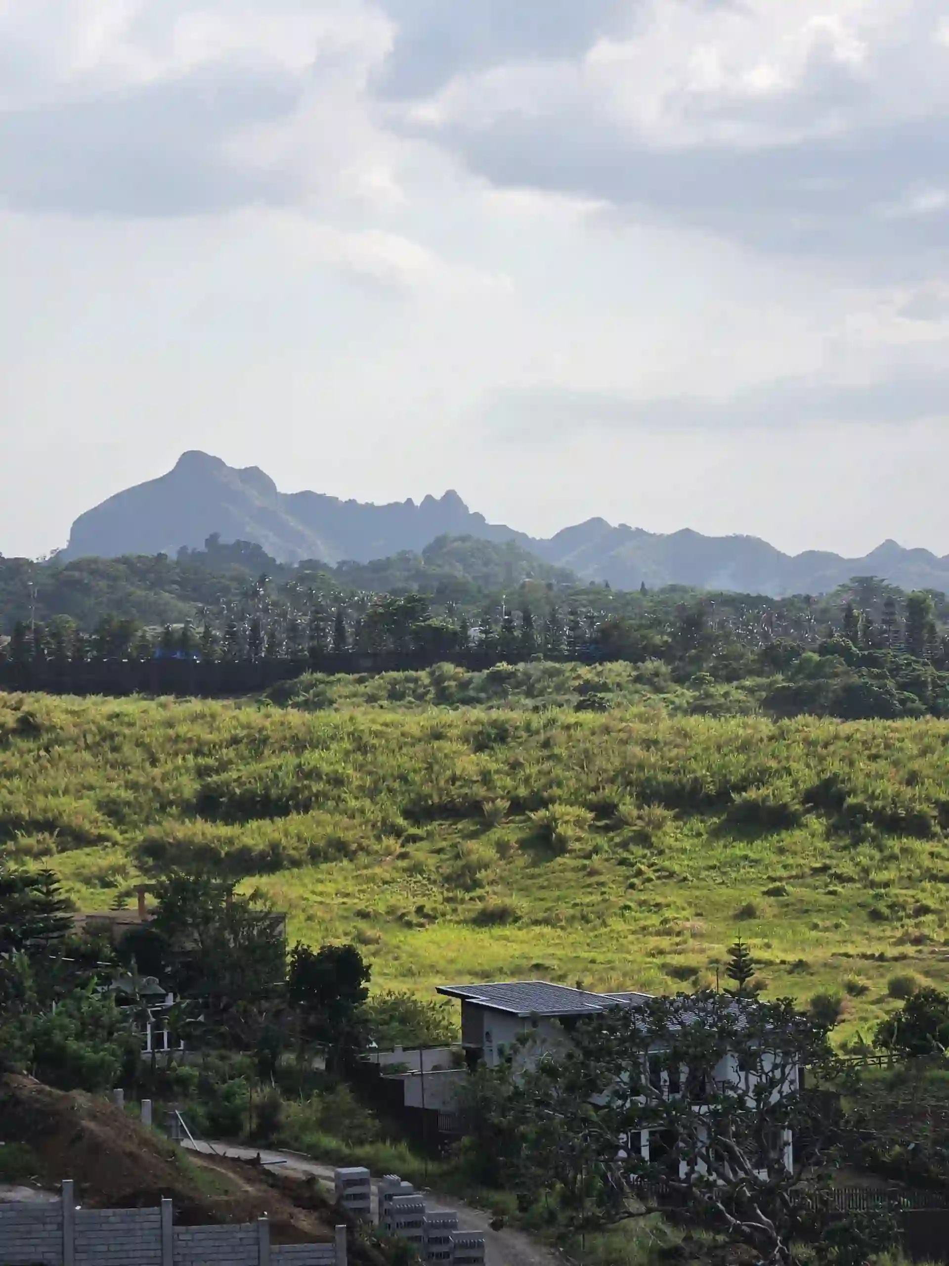 View of open farmland and grassy fields with houses in the foreground and mountain peaks along Amuyong Kaytitinga Road in Cavite under a cloudy sky