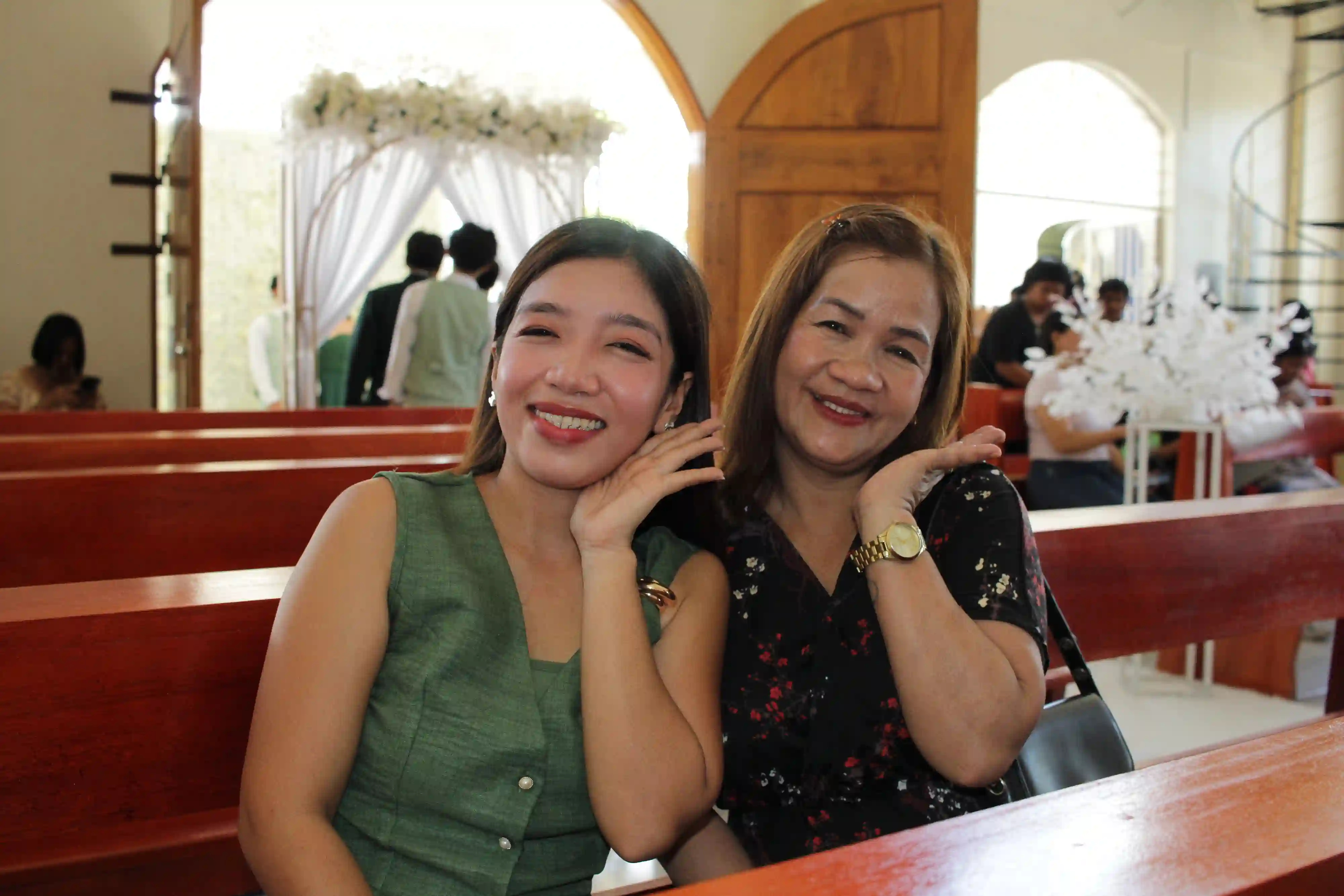 Two women smiling while seated on a wooden pew inside Our Lady of Fatima Mission Area in Socorro Oriental Mindoro with the floral entrance arch visible in the background