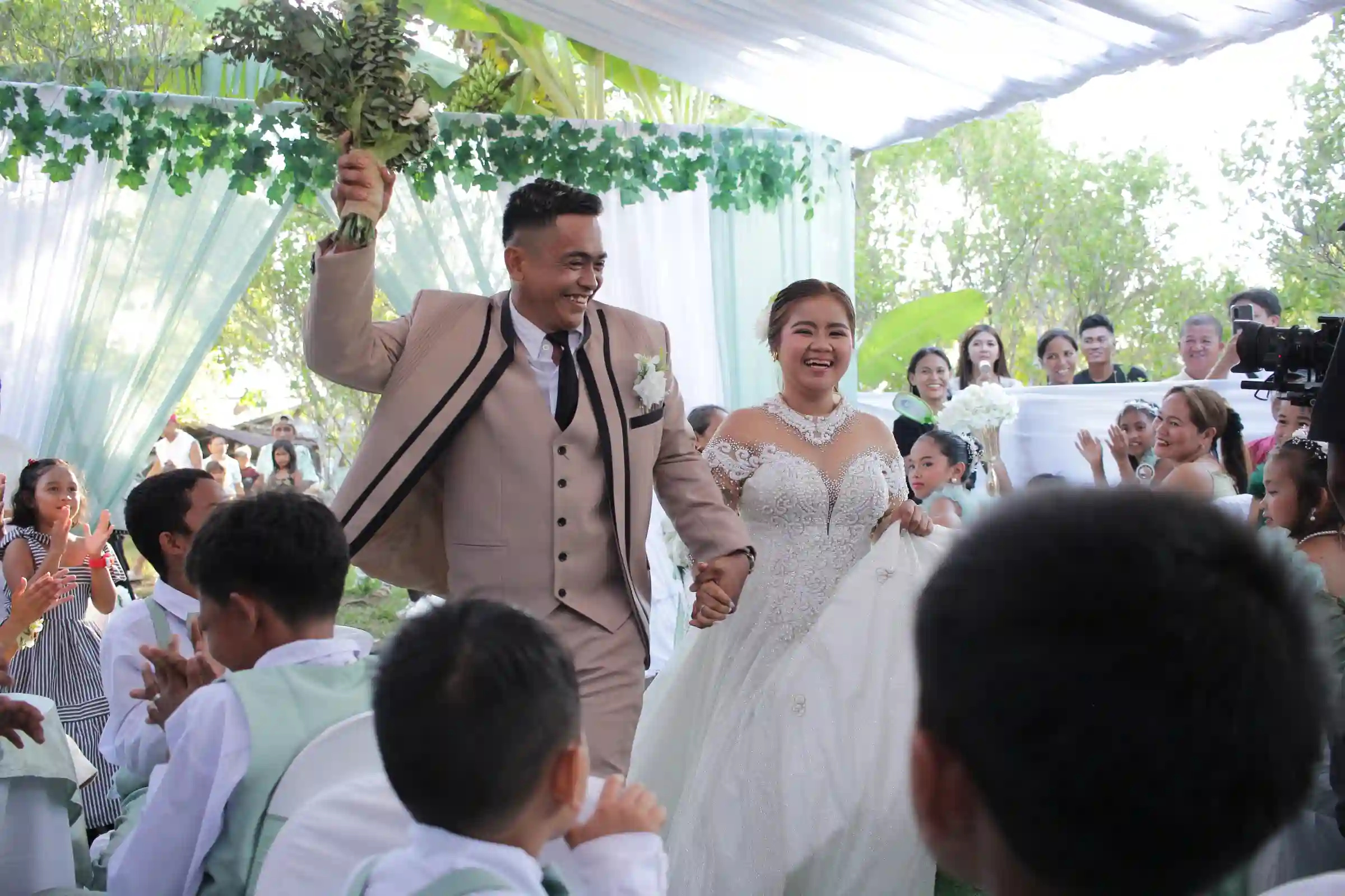 Newlyweds John Aries Almendras and Lyka Isabel Bastida exiting after their wedding ceremony with Aries raising the bouquet as guests applaud in Socorro Oriental Mindoro