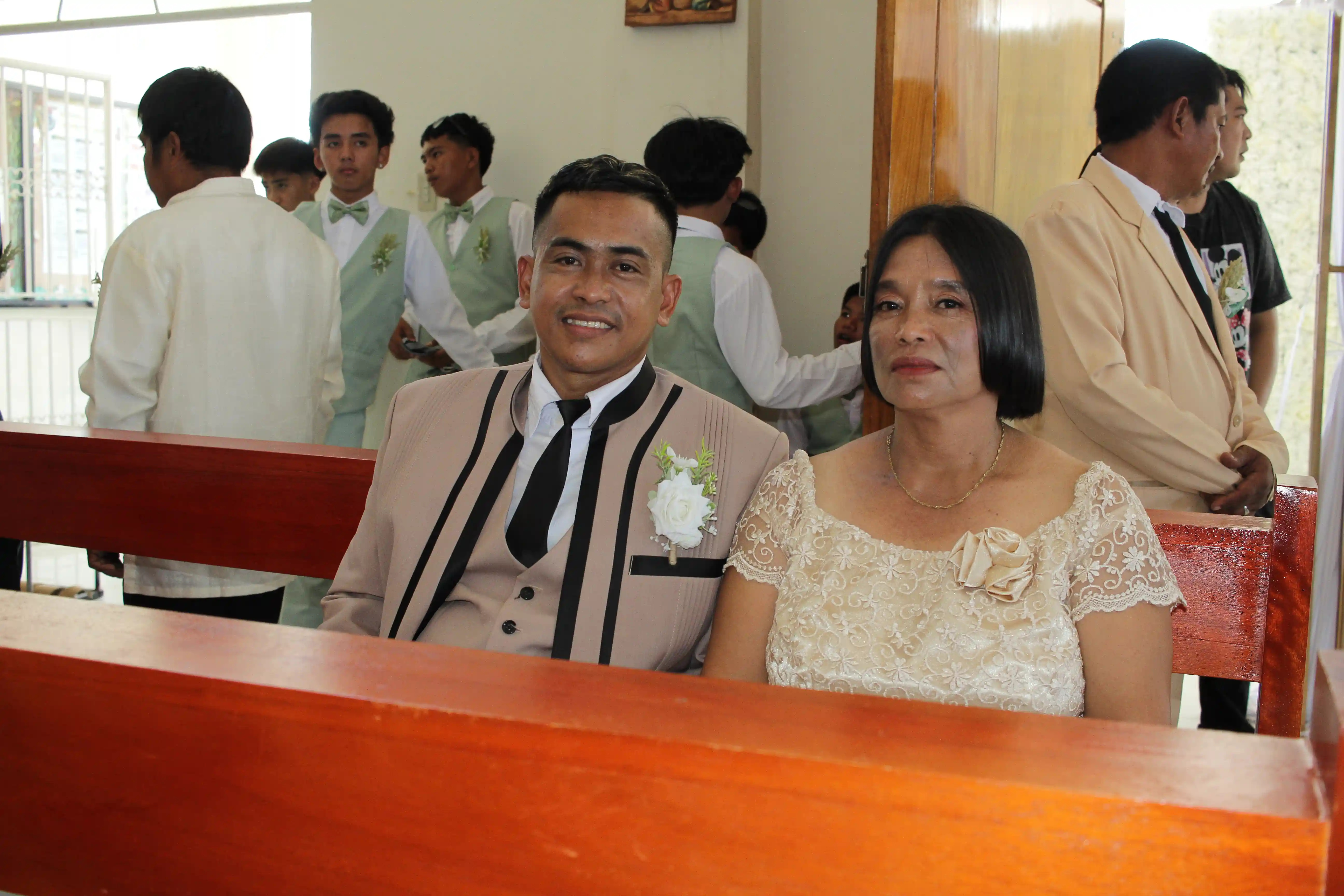 John Aries Almendras in champagne suit with boutonniere seated beside his mother in a cream lace blouse on a church pew before the wedding ceremony at Our Lady of Fatima Mission Area in Socorro Oriental Mindoro