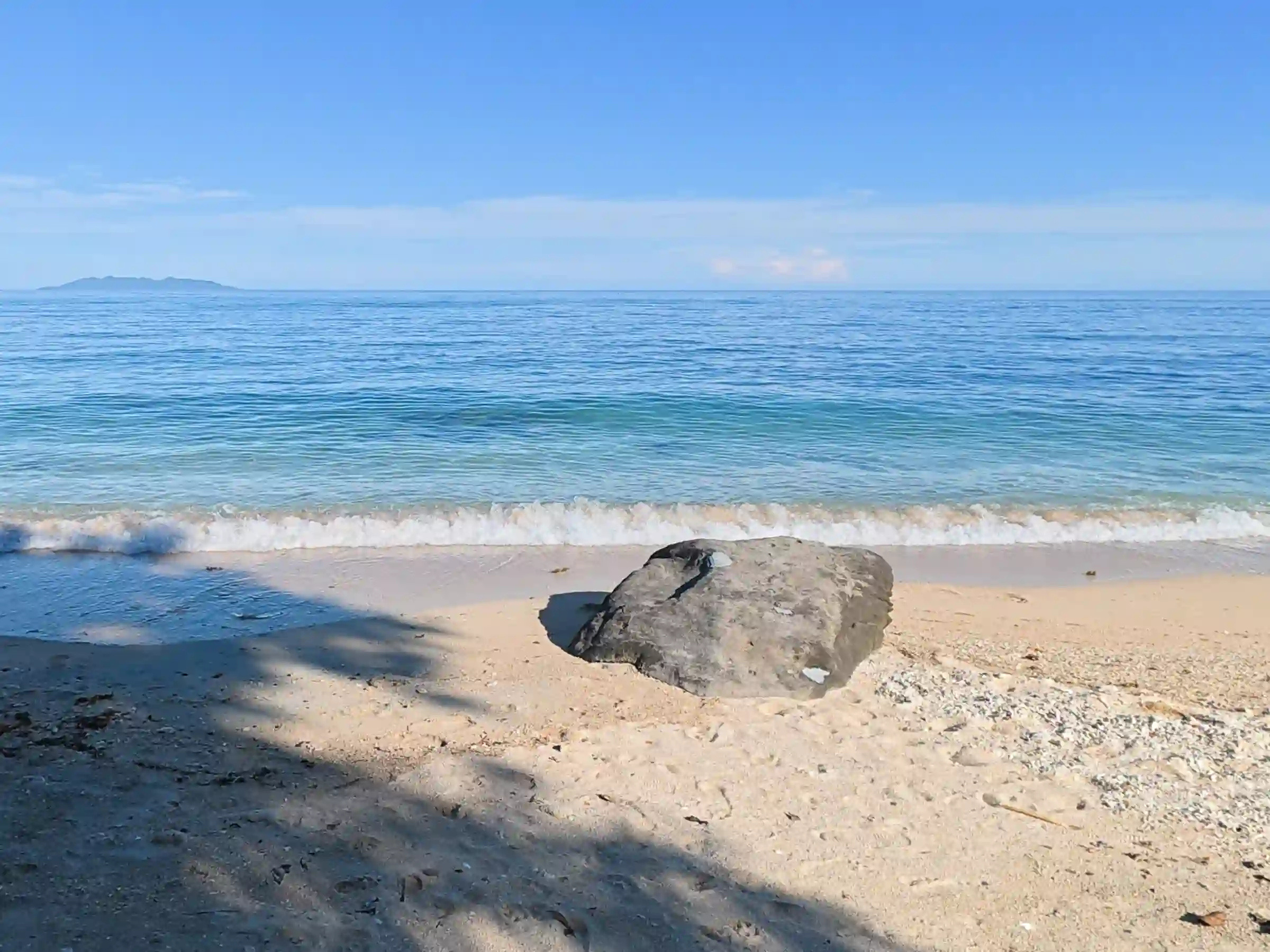 Single dark rock sitting on cream colored sand at the shoreline of Pili Beach in Pinamalayan Oriental Mindoro with Conception Island visible on the horizon