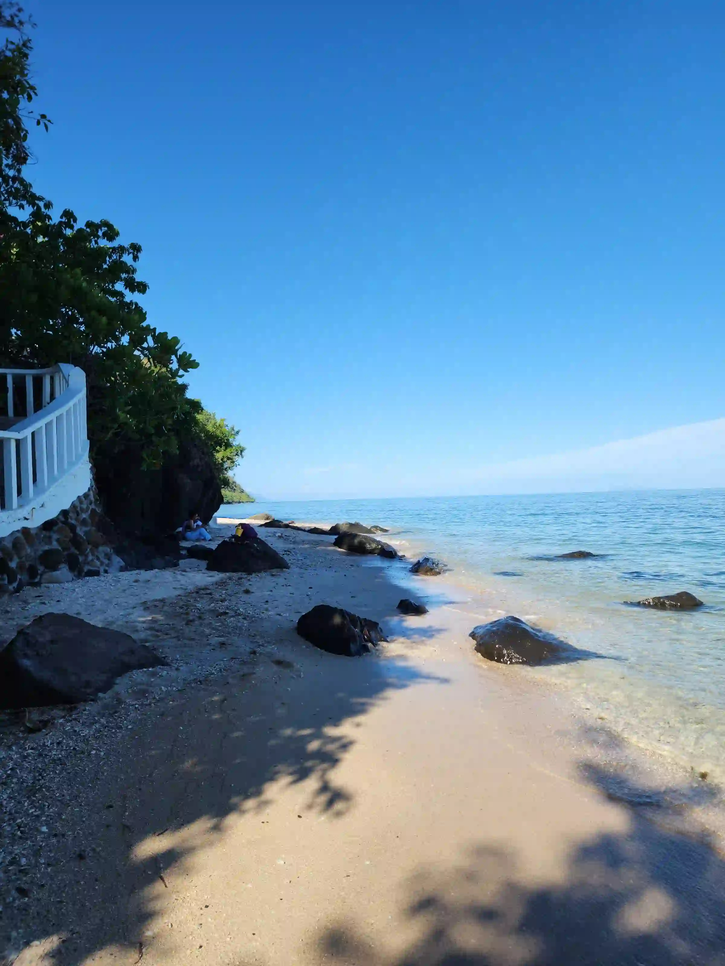 Rock scattered cream sand shoreline with two visitors resting near a resort railing at Pili Beach in Pinamalayan Oriental Mindoro