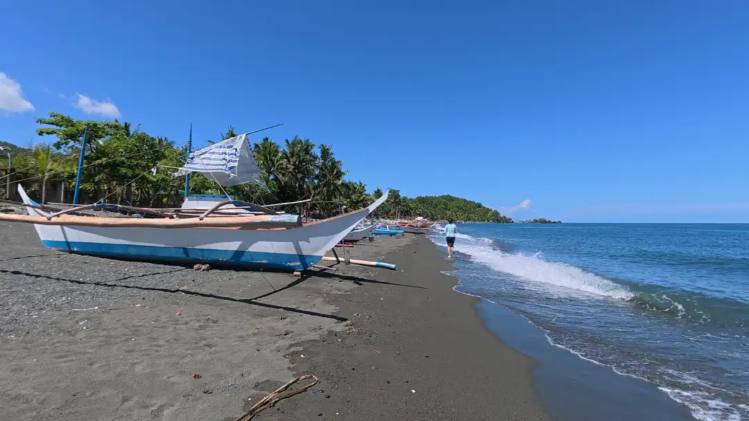 White and blue bangka fishing boats moored on dark volcanic sand at the far end of Sunrise Beach Resort Pili Pinamalayan Oriental Mindoro with a forested headland visible in the background