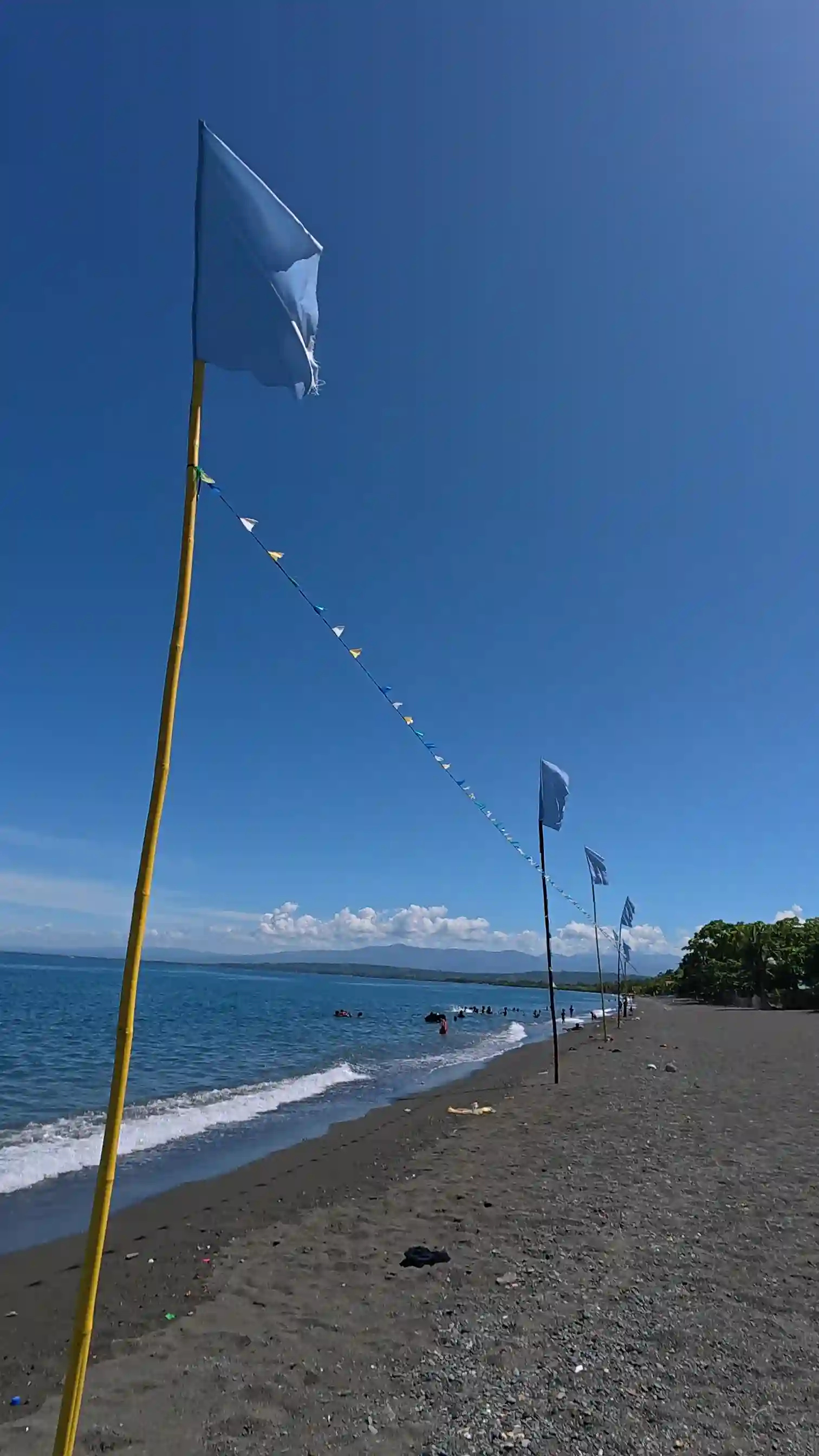 Row of blue flags on bamboo poles with colorful pennant bunting along the dark sand shoreline at Sunrise Beach Resort Pili Pinamalayan Oriental Mindoro mountains visible across the water